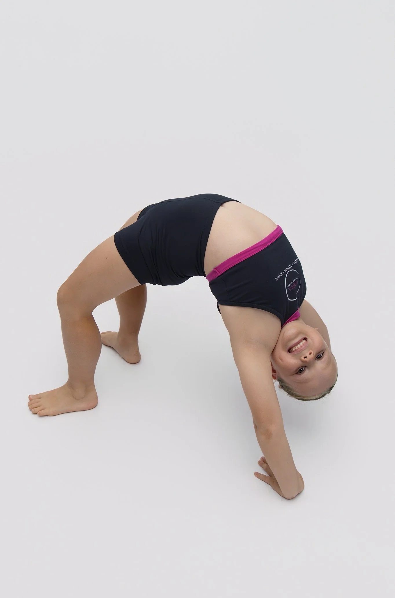 Young girl in black athletic shorts and pink waistband doing a yoga backbend on a white background, smiling.