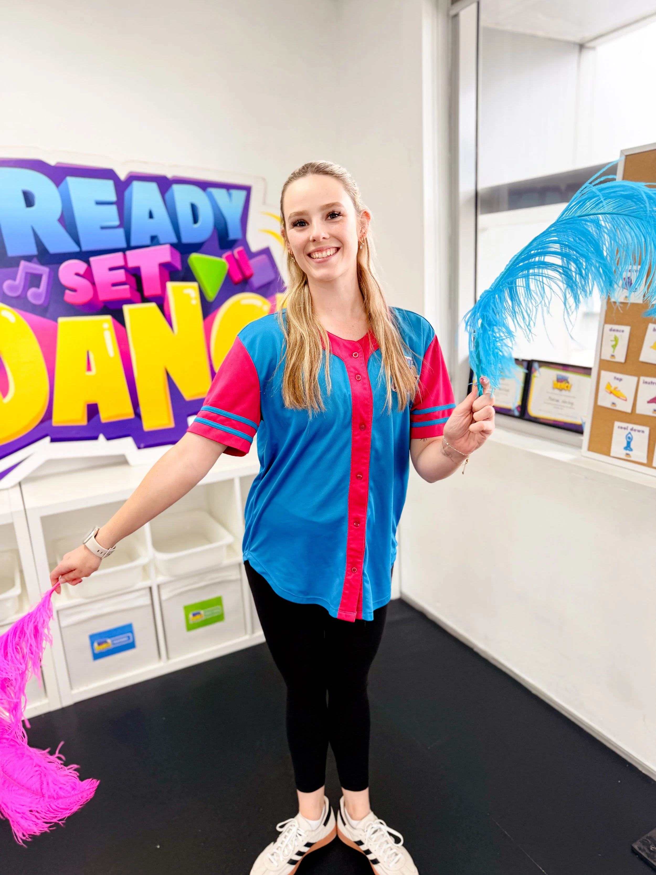 A young woman with blonde hair smiling, holding two bright pink and blue feathered boas, standing in front of a colorful sign that says 'Ready Set Dance' in a dance studio or classroom.