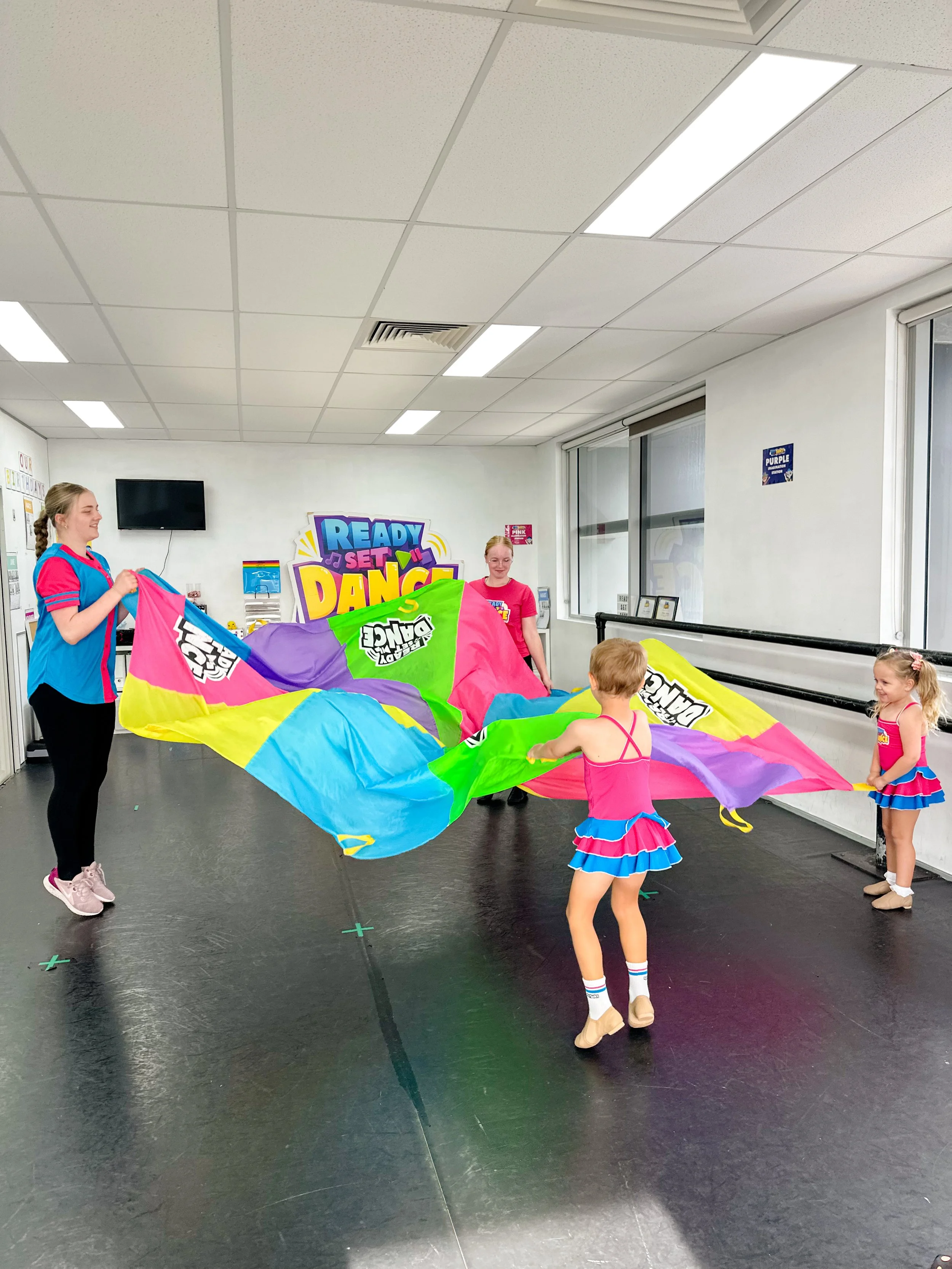 Children and two adults playing with a large colorful parachute in a dance studio.