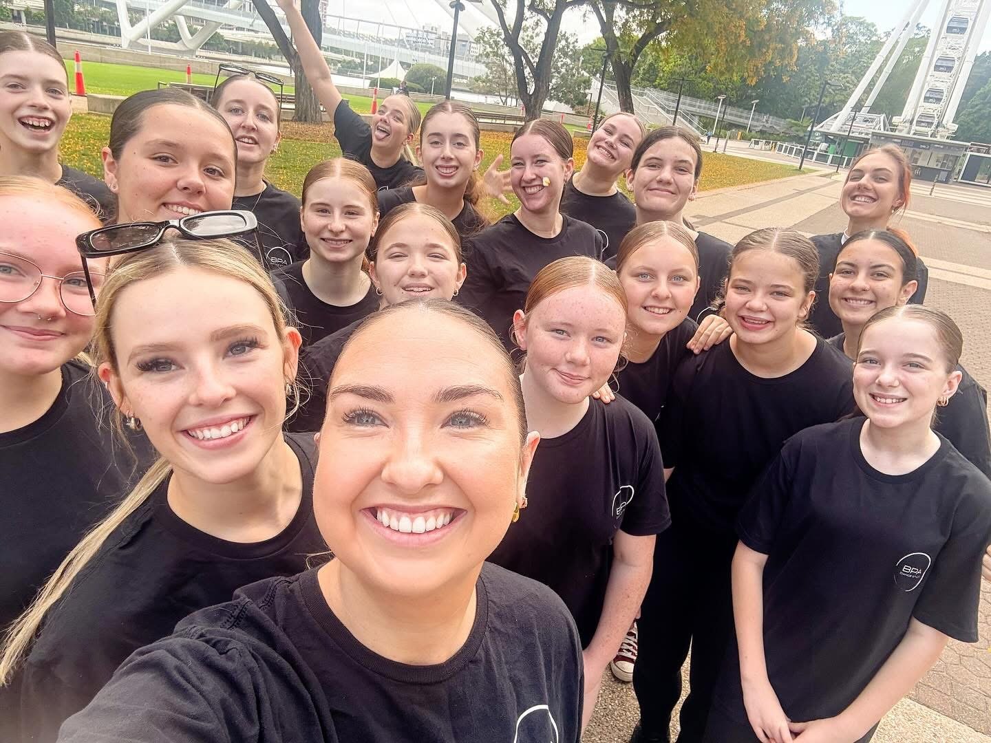 A group of young women in black shirts smiling and taking a selfie together outdoors near the riverfront, with trees and a Ferris wheel in the background.