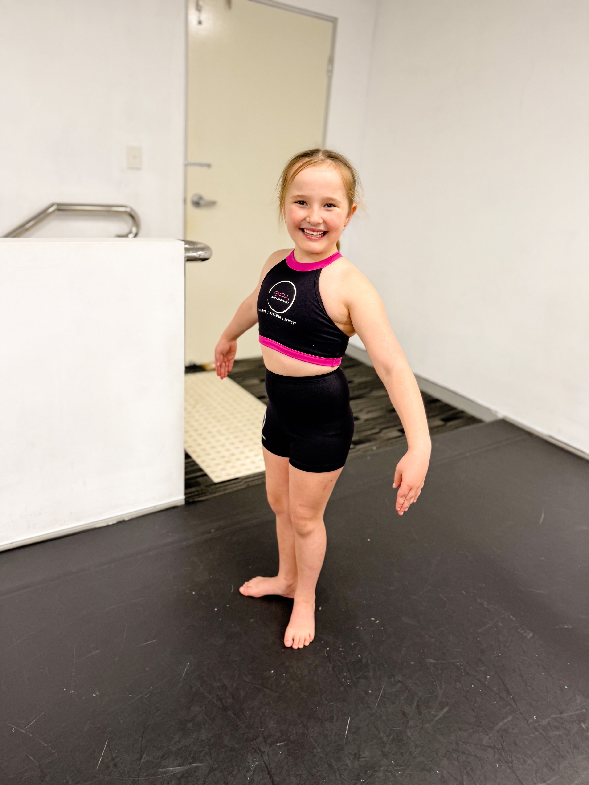 A young girl with blonde hair smiling and standing barefoot indoors, wearing a black athletic crop top with pink accents and black shorts.