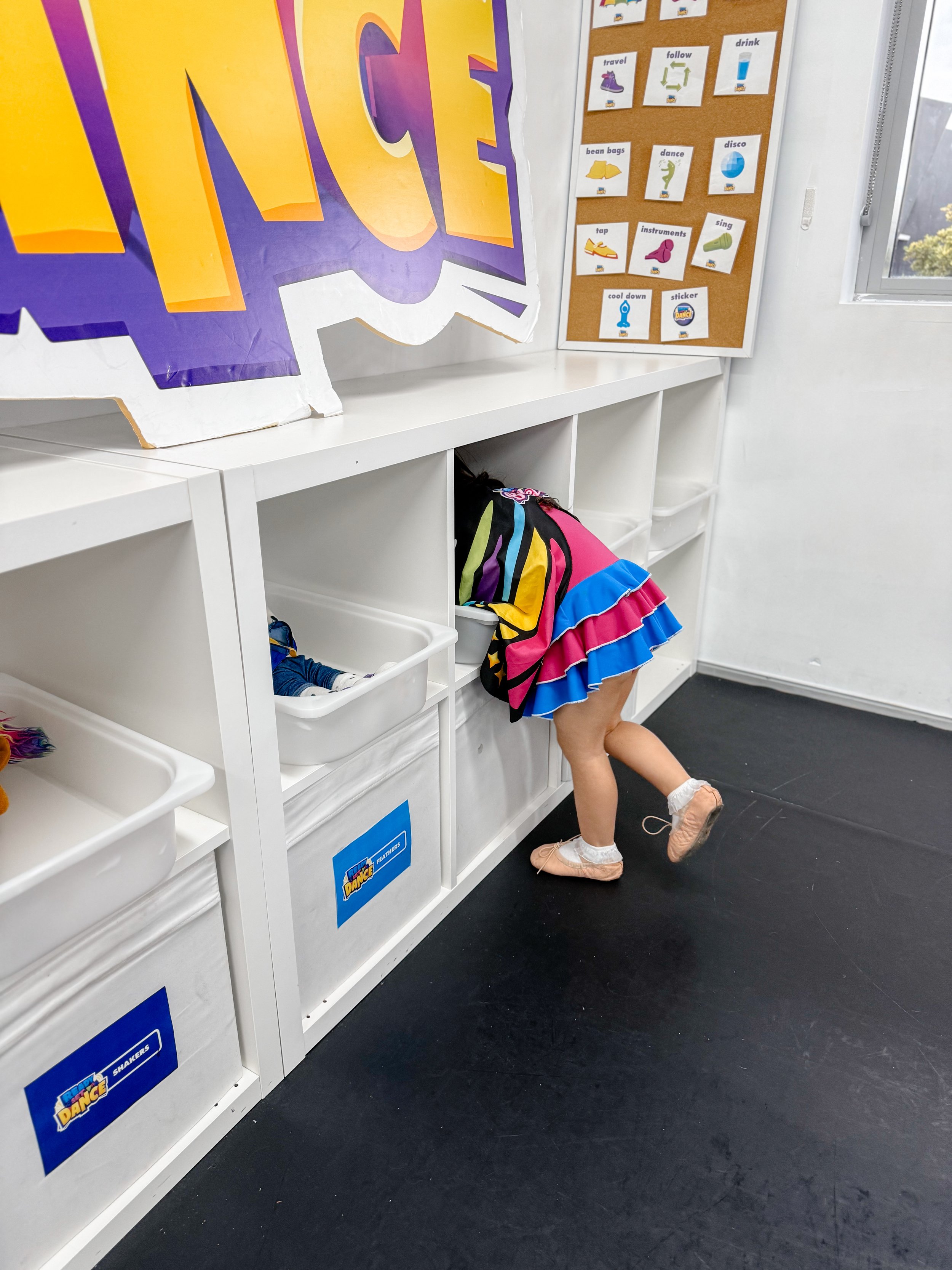 A young girl in a colorful dress with ruffles is bent over and searching inside a cubby bin at a white storage unit in a classroom. The classroom has posters with icons and words on the wall, and a partially visible large colorful sign.
