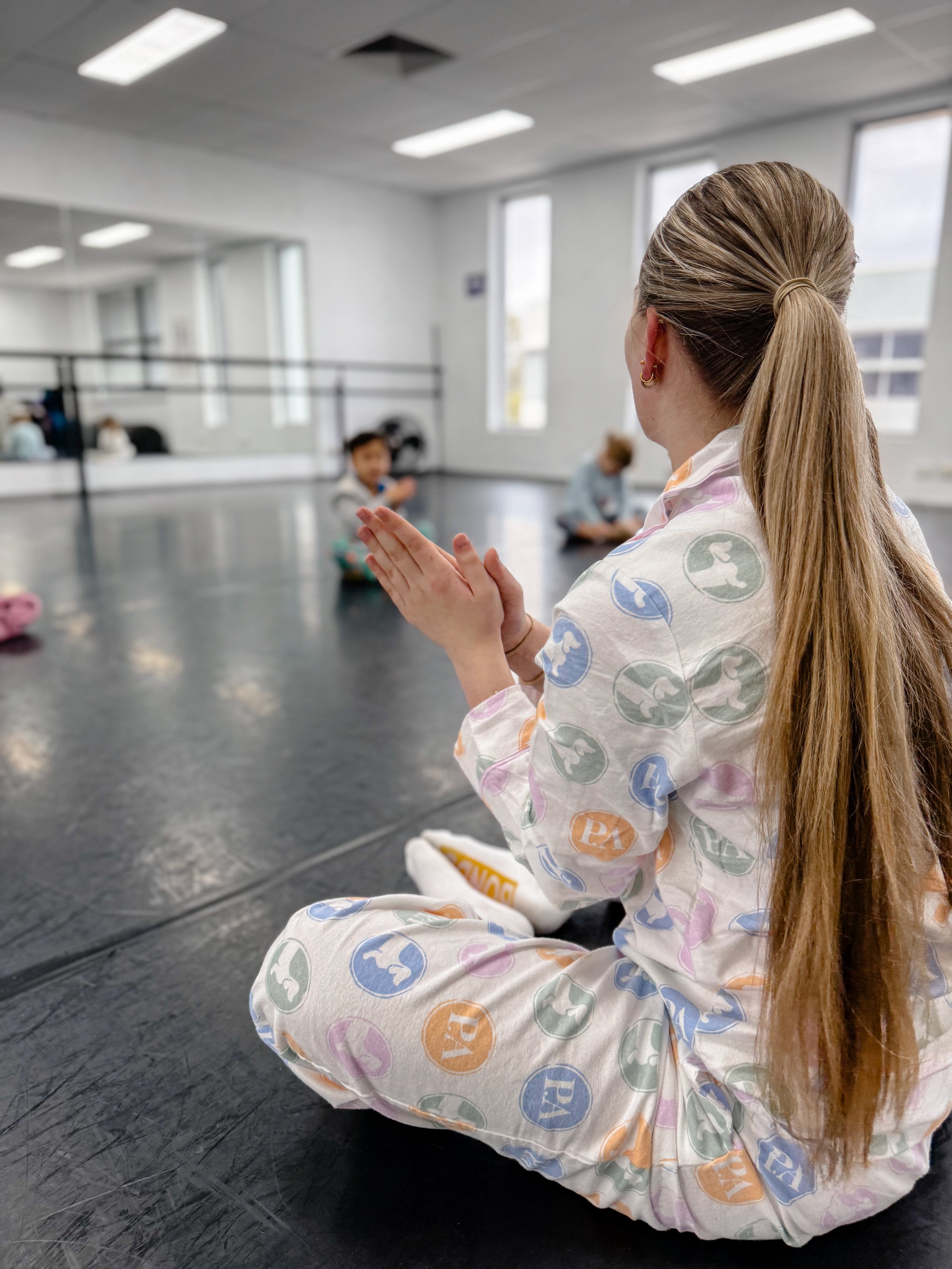 A woman with long hair in a ponytail, wearing colorful pajamas, sits cross-legged on a black floor in a dance studio, clapping her hands, while children practice seated on the floor in the background.