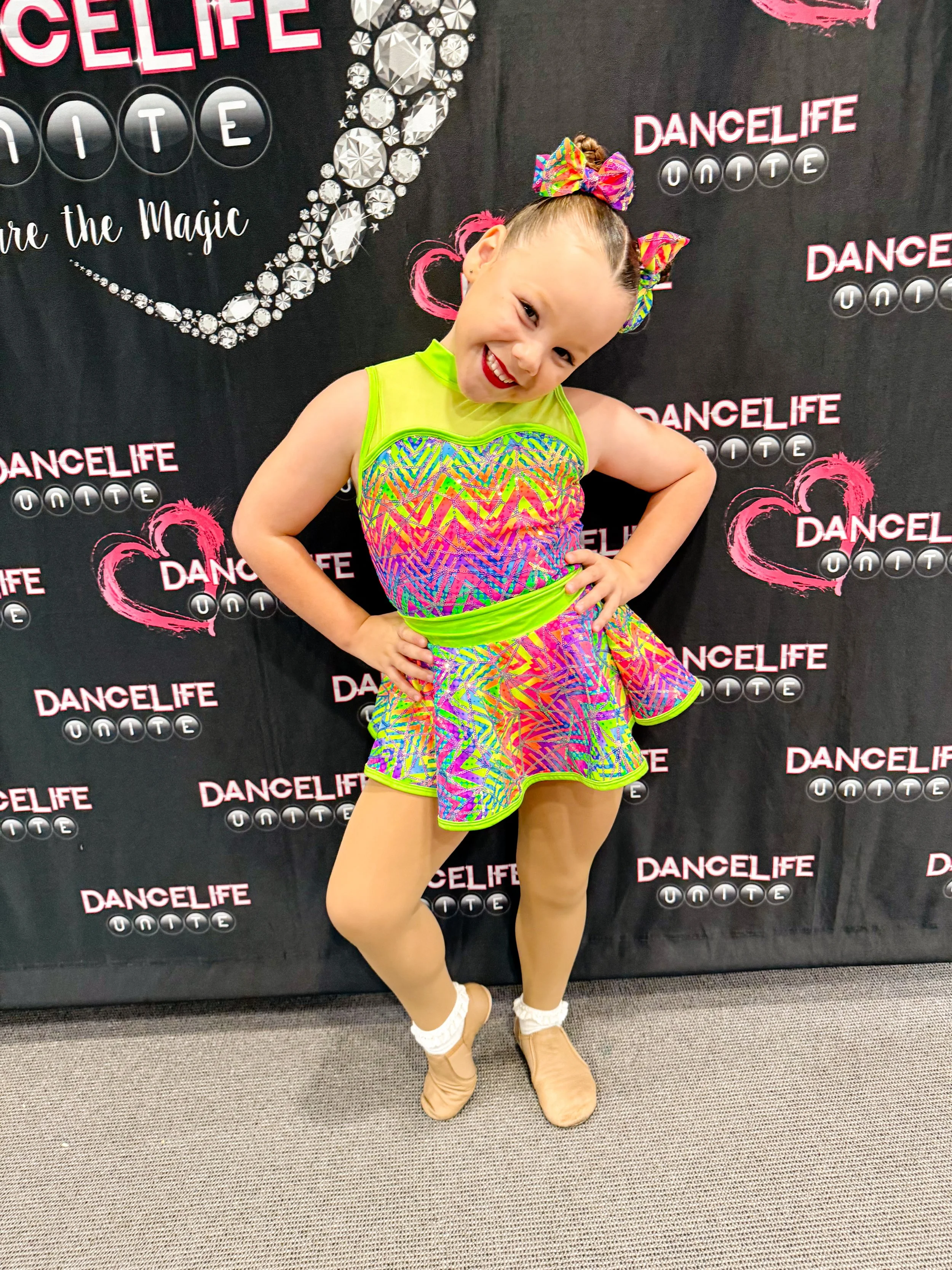 A young girl dressed in a colorful, neon dance costume with a tutu, beige dance shoes, and a matching headband, poses cheerfully in front of a black backdrop with pink and white lettering that reads "Dancelife Unite."