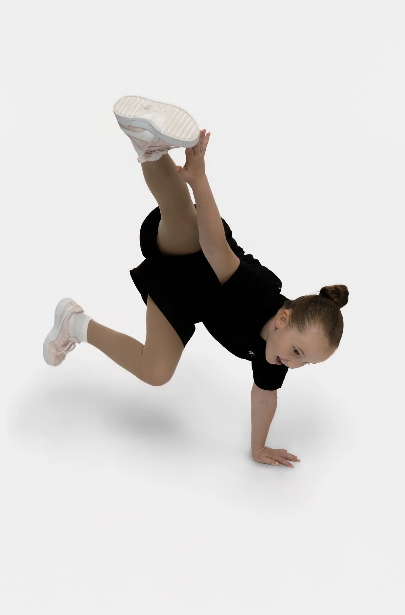 A young woman performing a breakdancing move on a white background, balancing on one hand with one leg raised and holding her other leg with her hand.