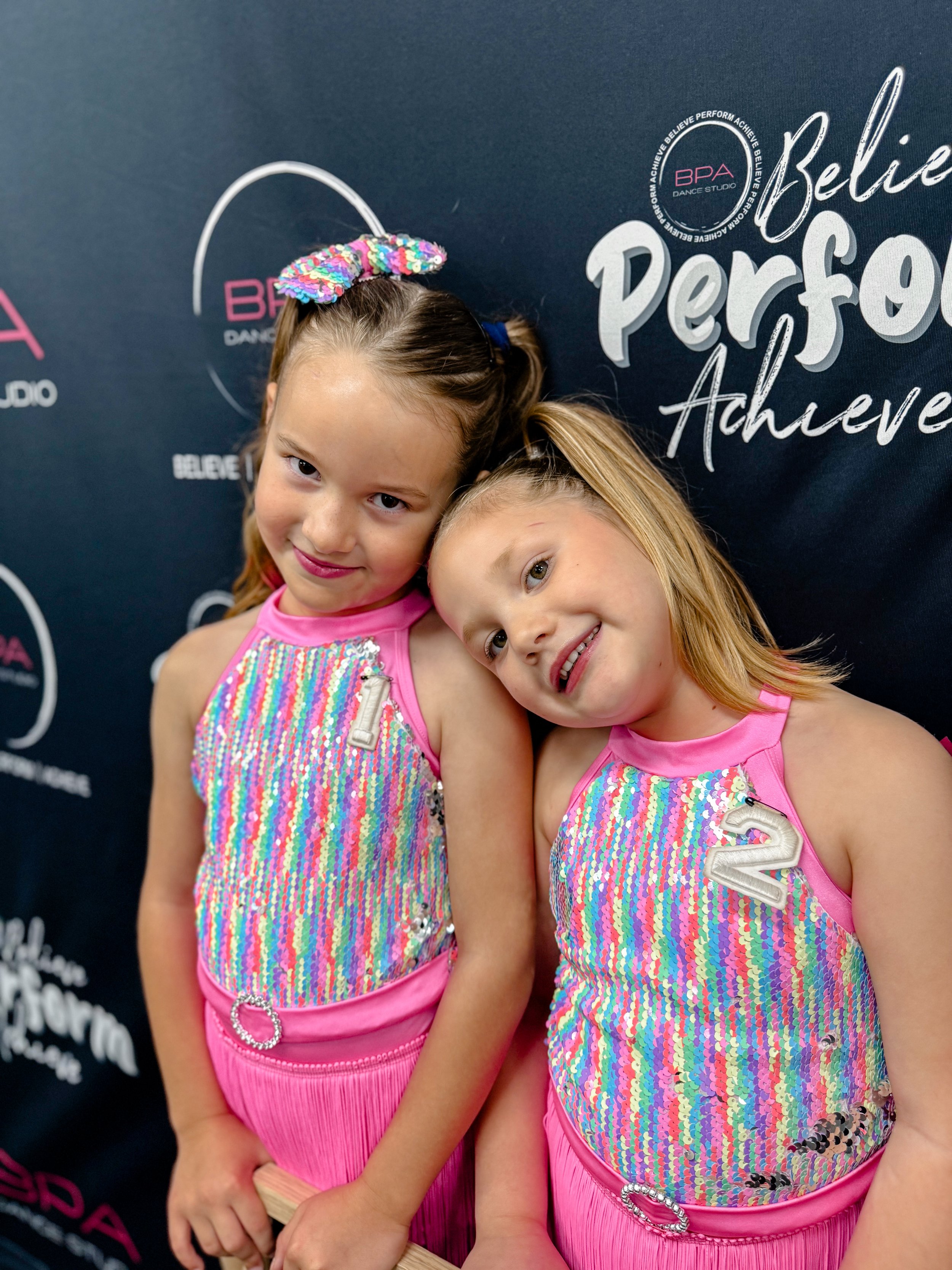Two young girls wearing matching sparkly pink dance costumes with numbers '1' and '2' on the chest, standing close together with their heads touching, smiling, in front of a backdrop with motivational words and logos.