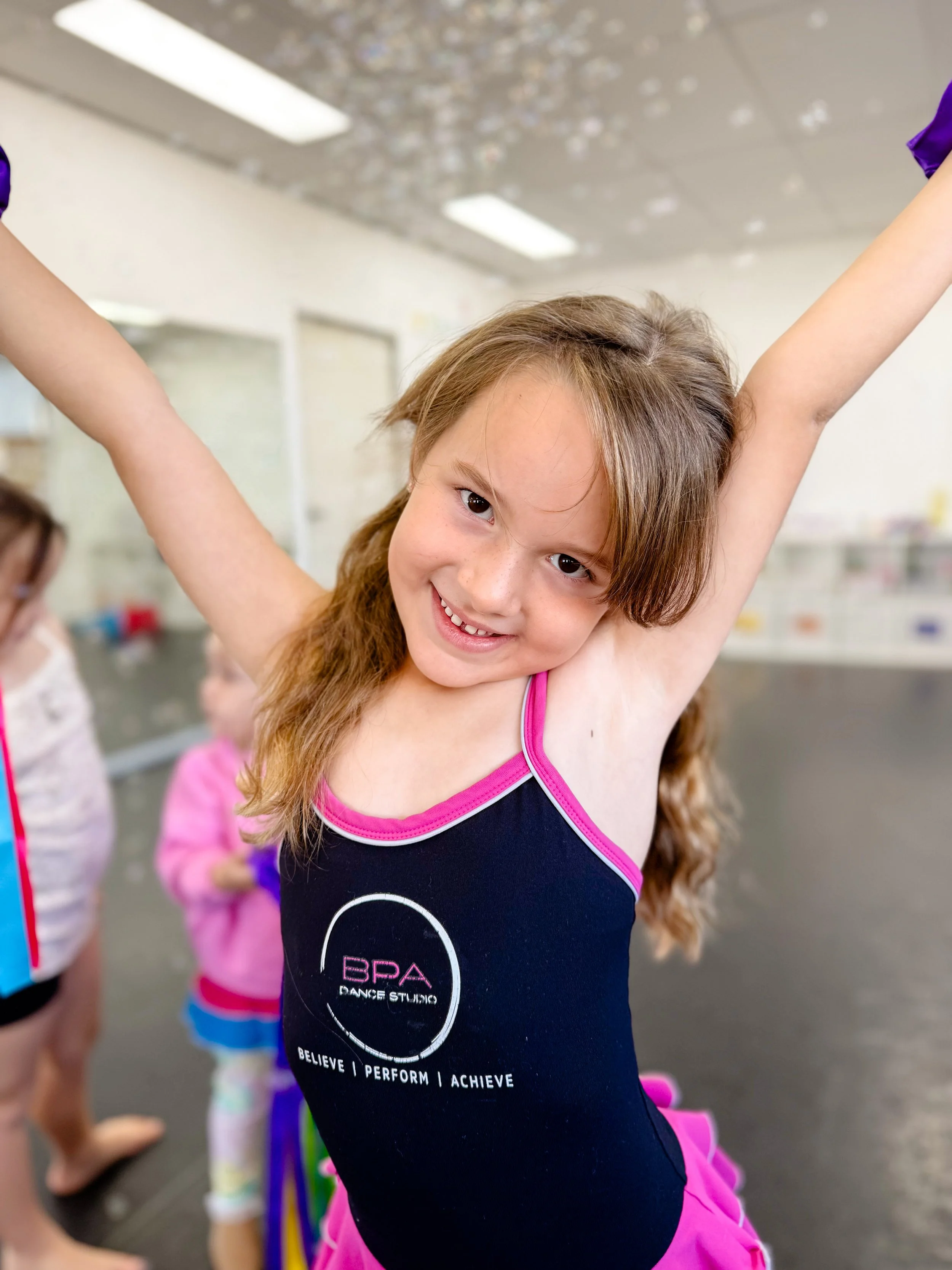 Young girl with long hair in a dance studio, smiling with arms raised, wearing a black and pink dance uniform with the logo 'BPA Dance Studio'.