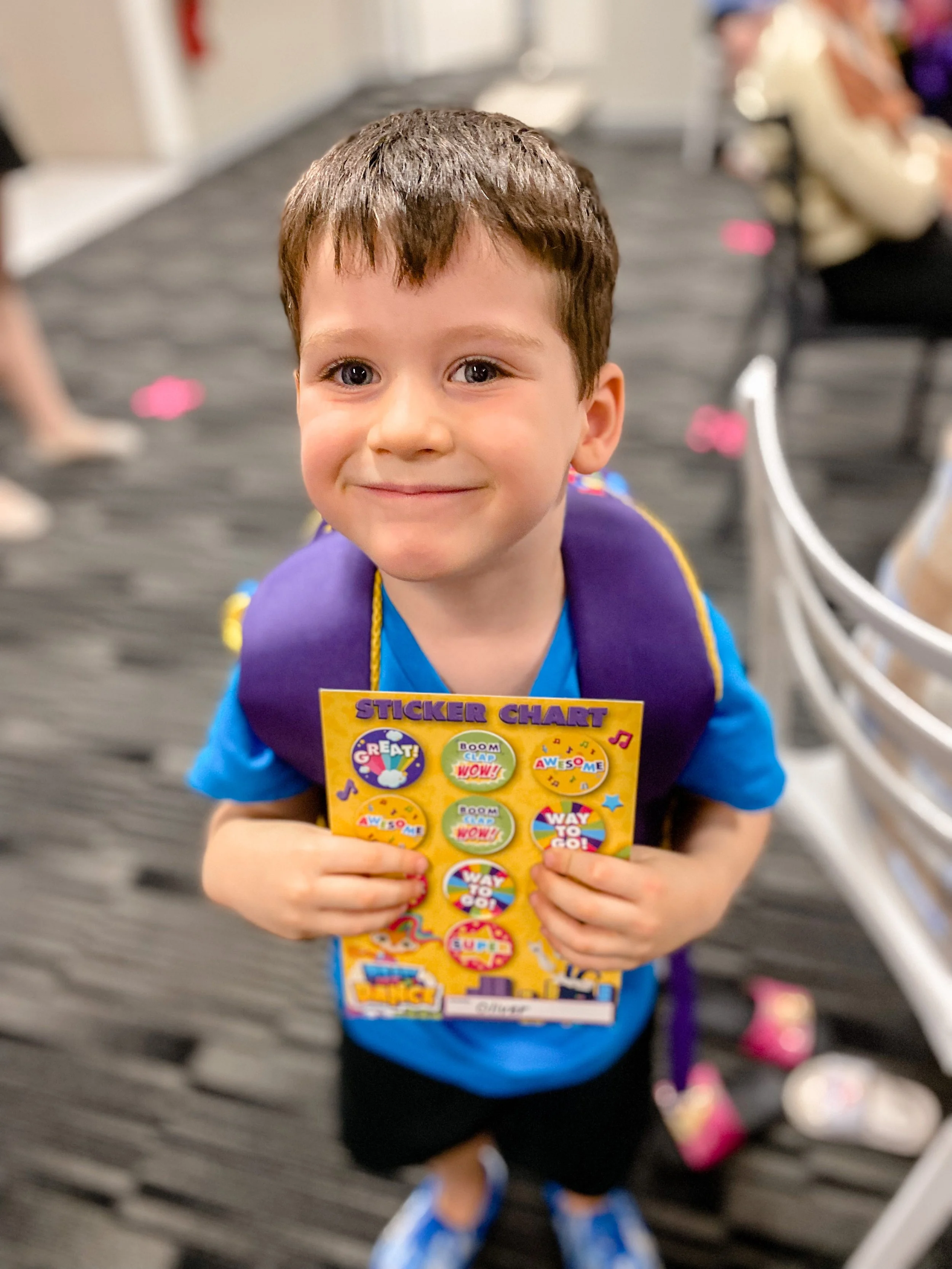 Young boy smiling and holding a colorful sticker chart, standing indoors on a gray and black striped carpet with some pink and white items on the floor.