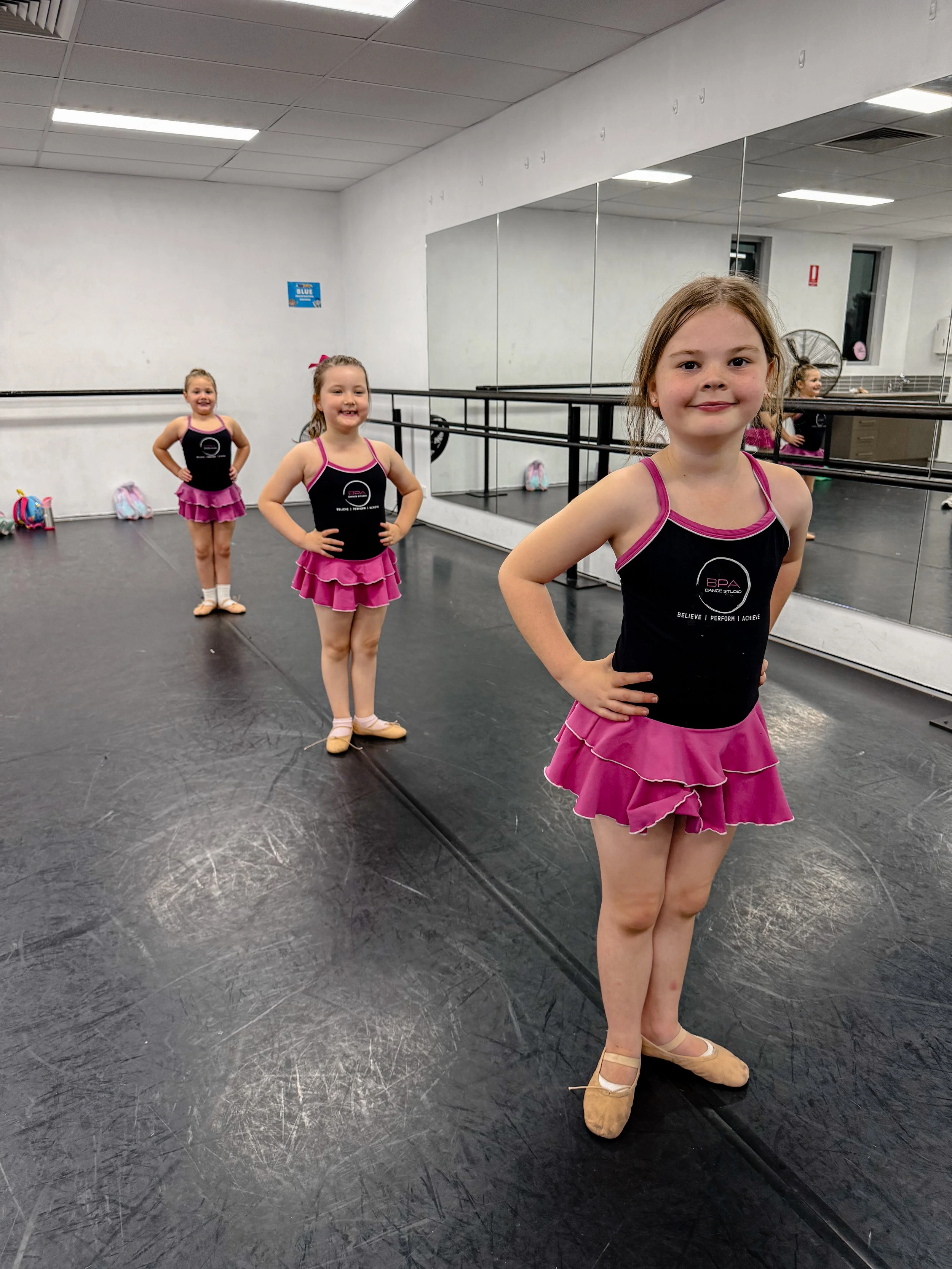 Four young girls in ballet costumes standing in order in a dance studio with mirrored walls and ballet floors, smiling and posing with their hands on their hips.