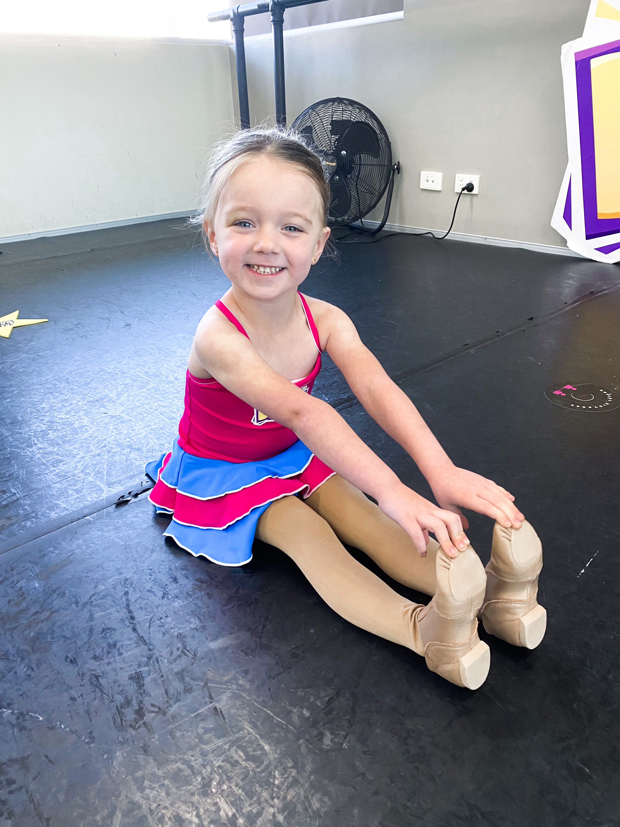 A young girl in a pink and blue cheerleading outfit sitting on the floor stretching, with a big smile, in a room with black flooring, a fan, and a large colorful sign
