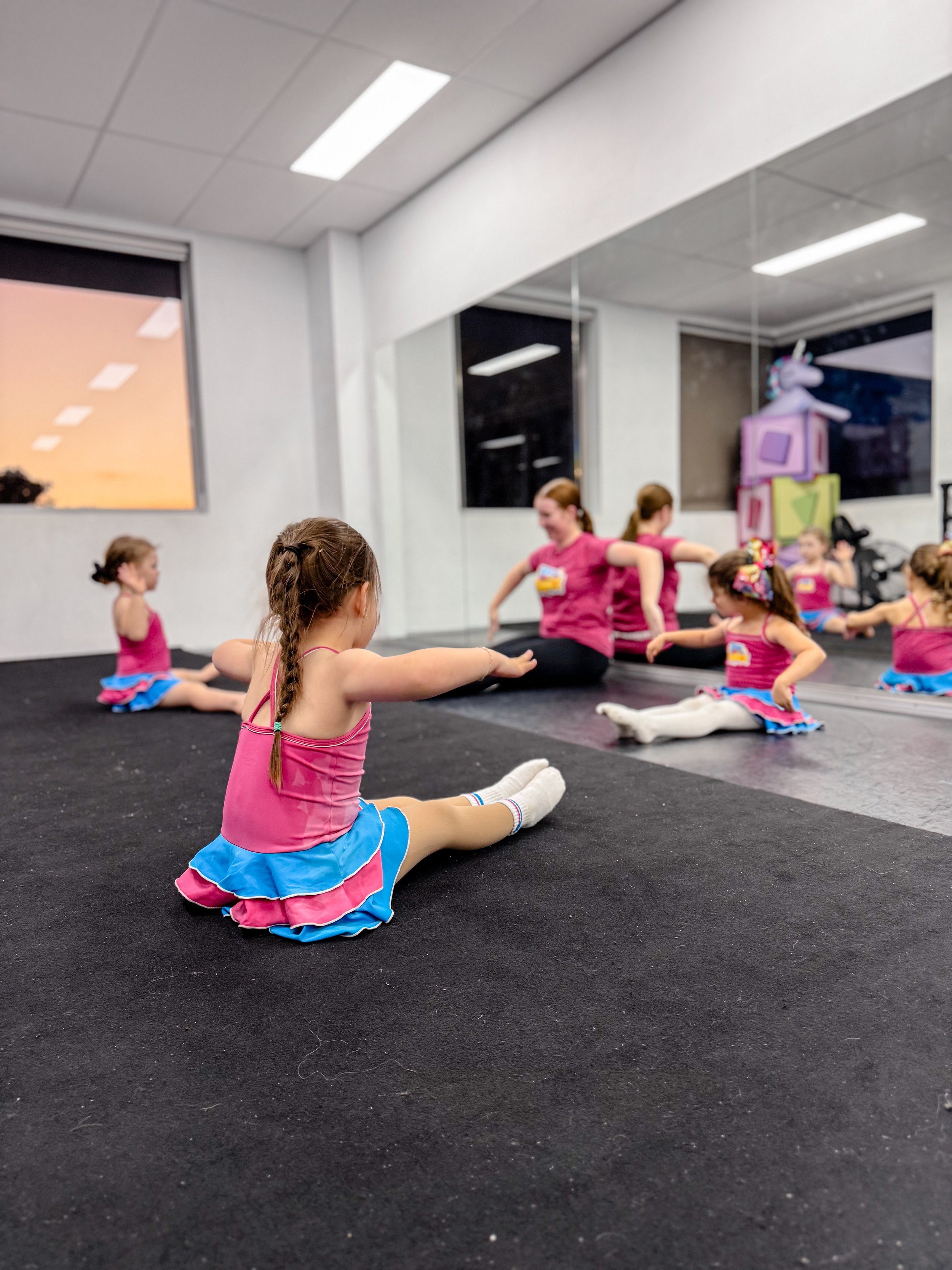 Young girls in colorful dance costumes sitting on the floor during a dance class, with a mirrored wall and windows showing a sunset
