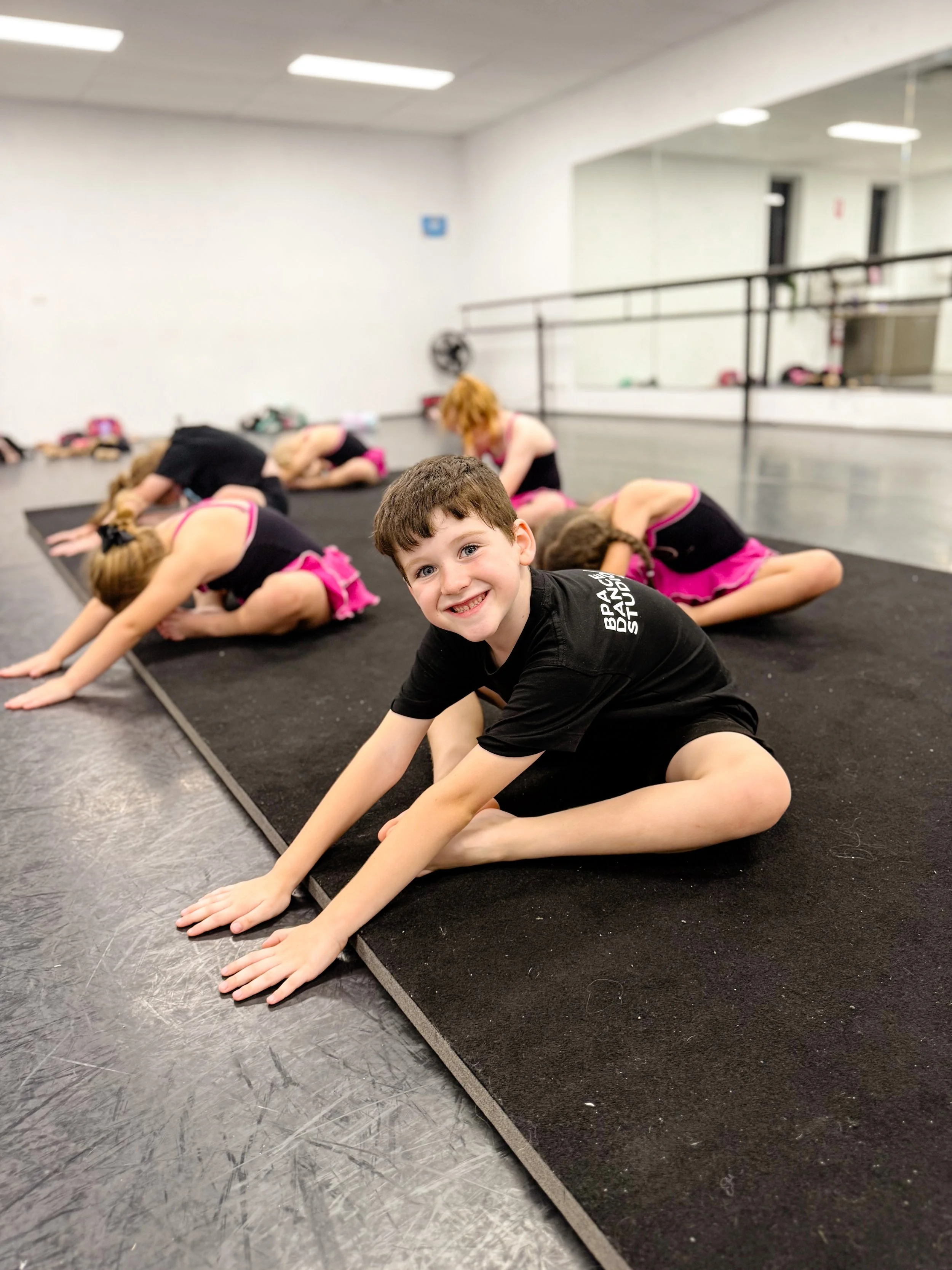 Young boy with short brown hair smiling while sitting in a split stretch on a black yoga mat in a dance studio, with several girls in black and pink dance outfits doing stretches in the background.