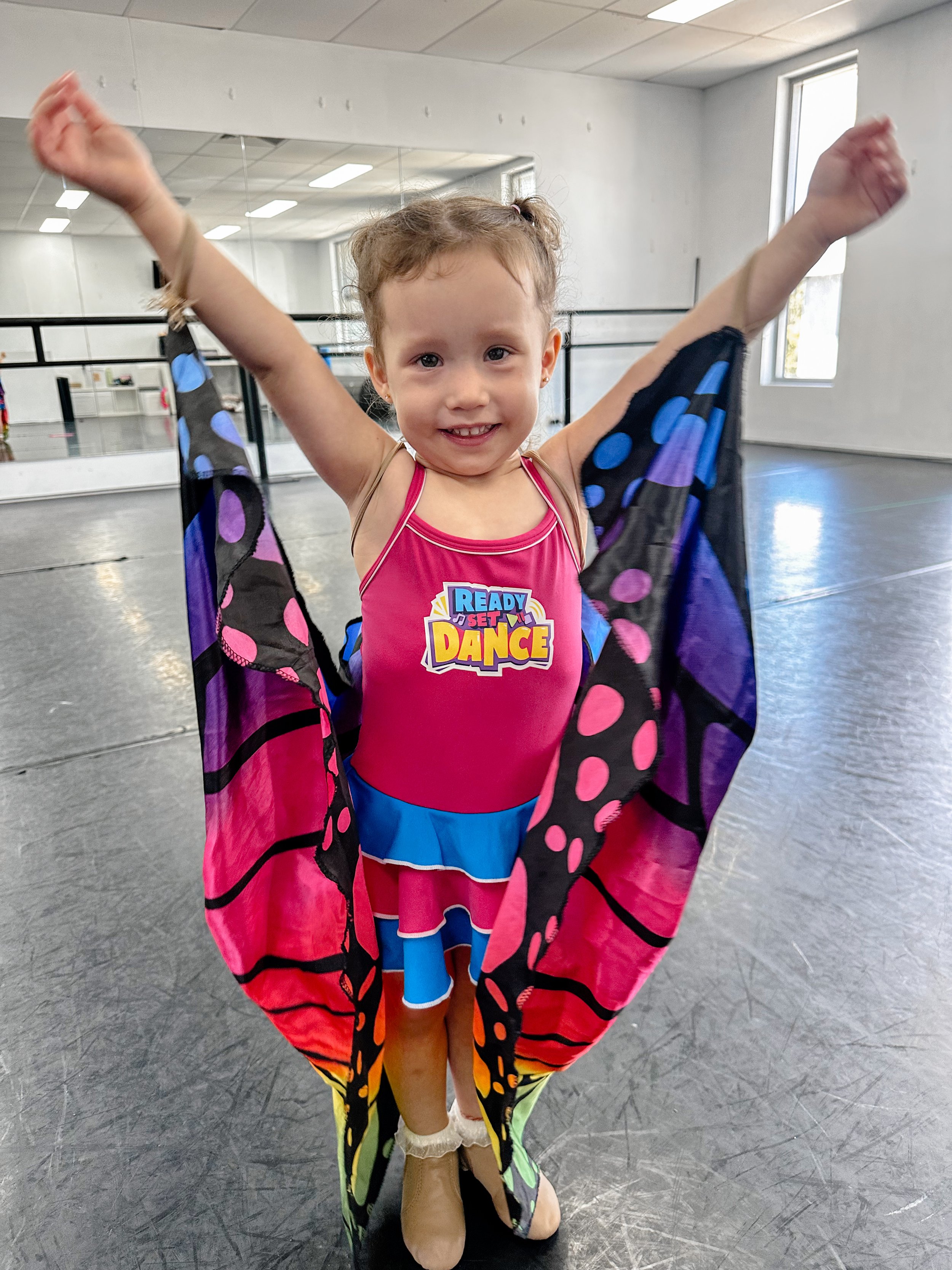 Young girl in dance costume with butterfly wings, smiling with arms raised in a dance studio.