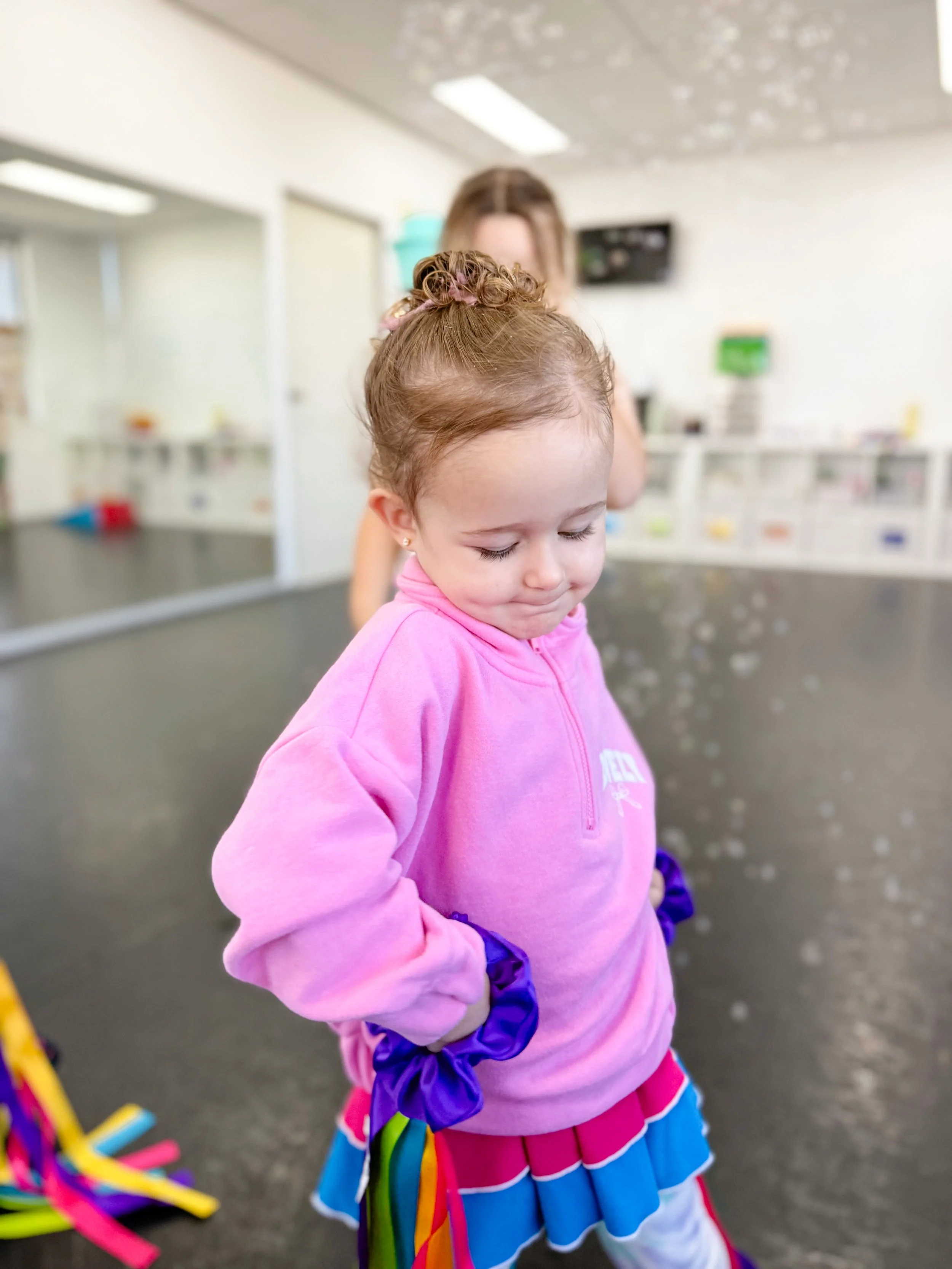 A young girl with a curly bun wearing a pink hoodie and a colorful layered skirt, standing with her hands on her hips in an indoor play area.
