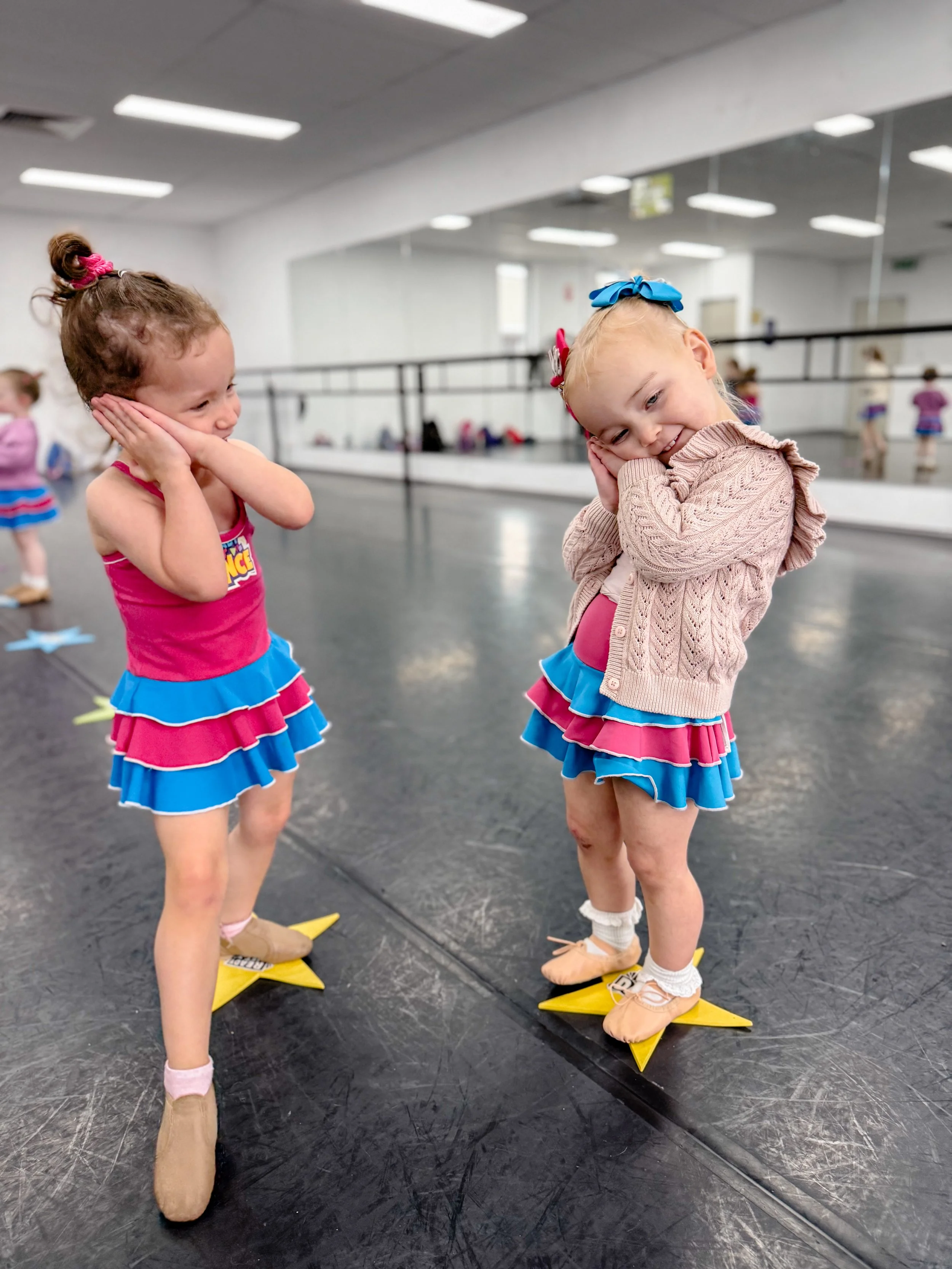 Two young girls in pink and blue dance costumes with ruffled skirts standing on yellow star-shaped floor markers in a dance studio, covering their ears and smiling.