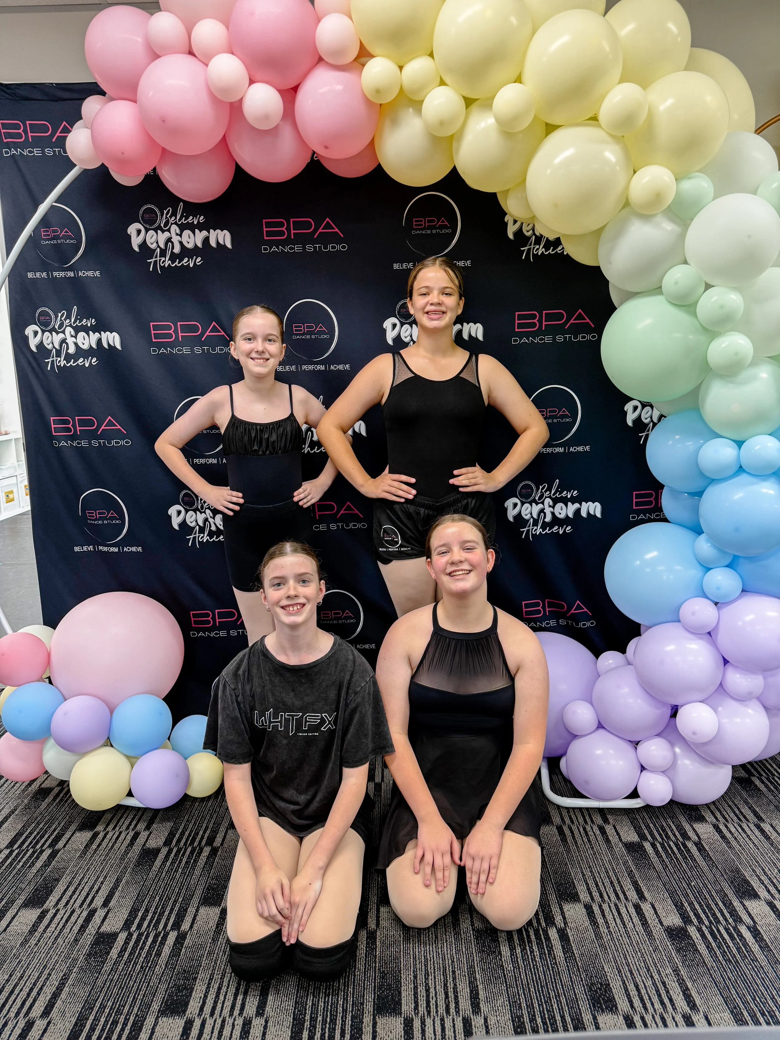 Four young female dancers in dance costumes pose in front of a decorated backdrop with balloons at a dance studio event.