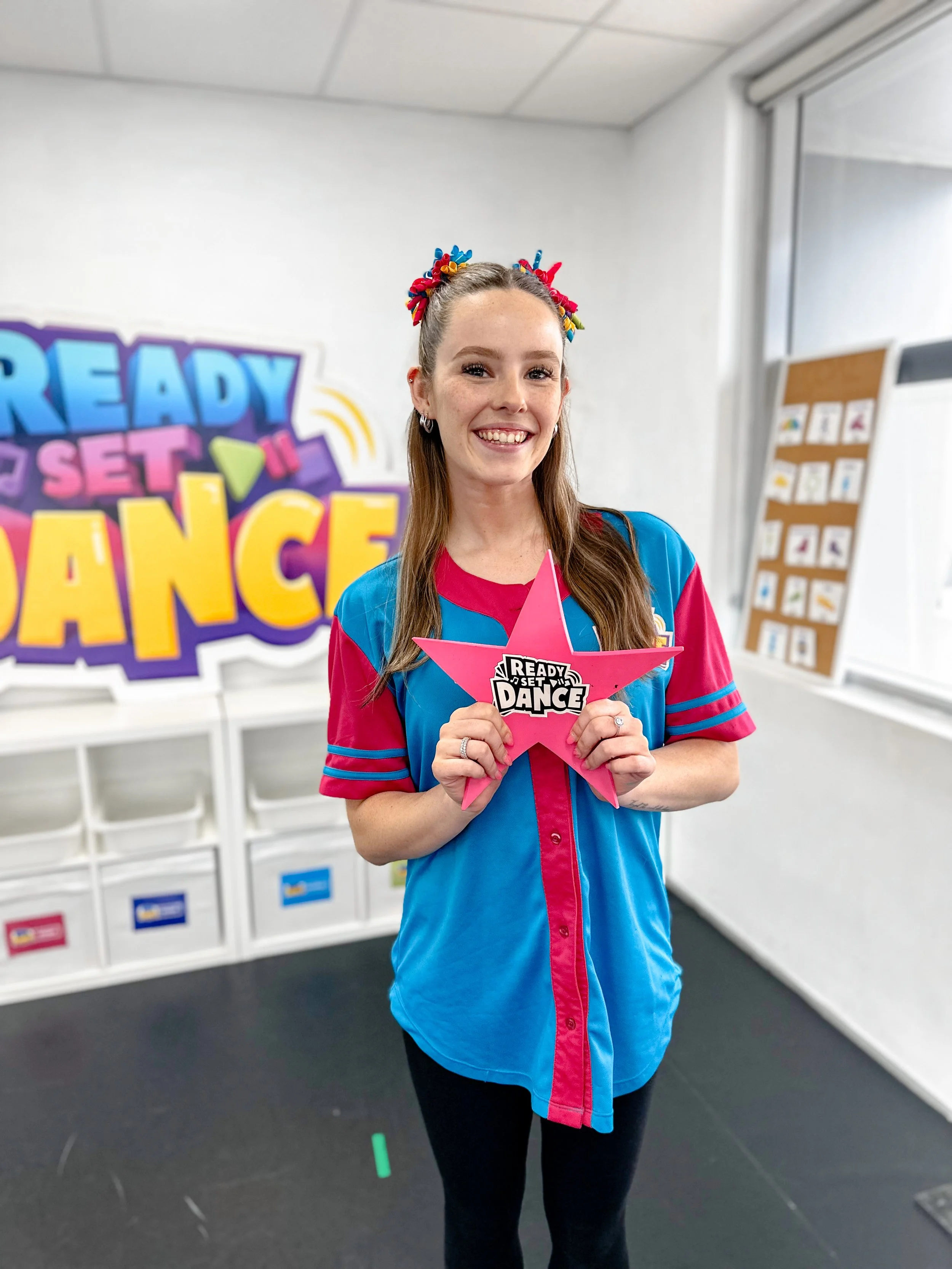 A young woman with long brown hair smiling, holding a pink star-shaped sign that says 'Ready, Set, Dance' in a dance studio with colorful decorations and a large sign on the wall that also says 'Ready, Set, Dance'