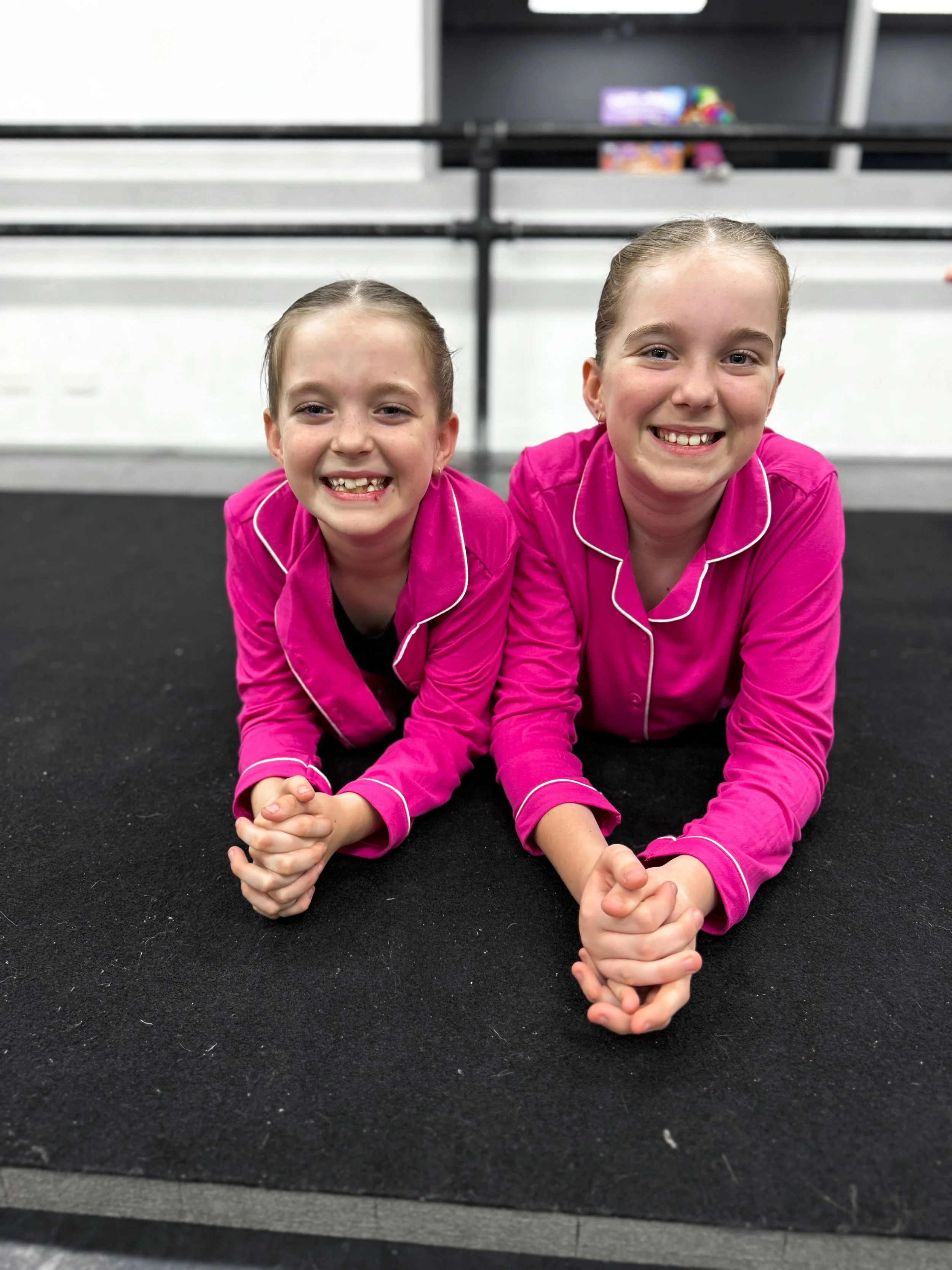 Two young girls lying on their stomachs on a black gym mat, smiling at the camera, wearing matching pink pajamas with white piping, in a gymnastics or gym setting.