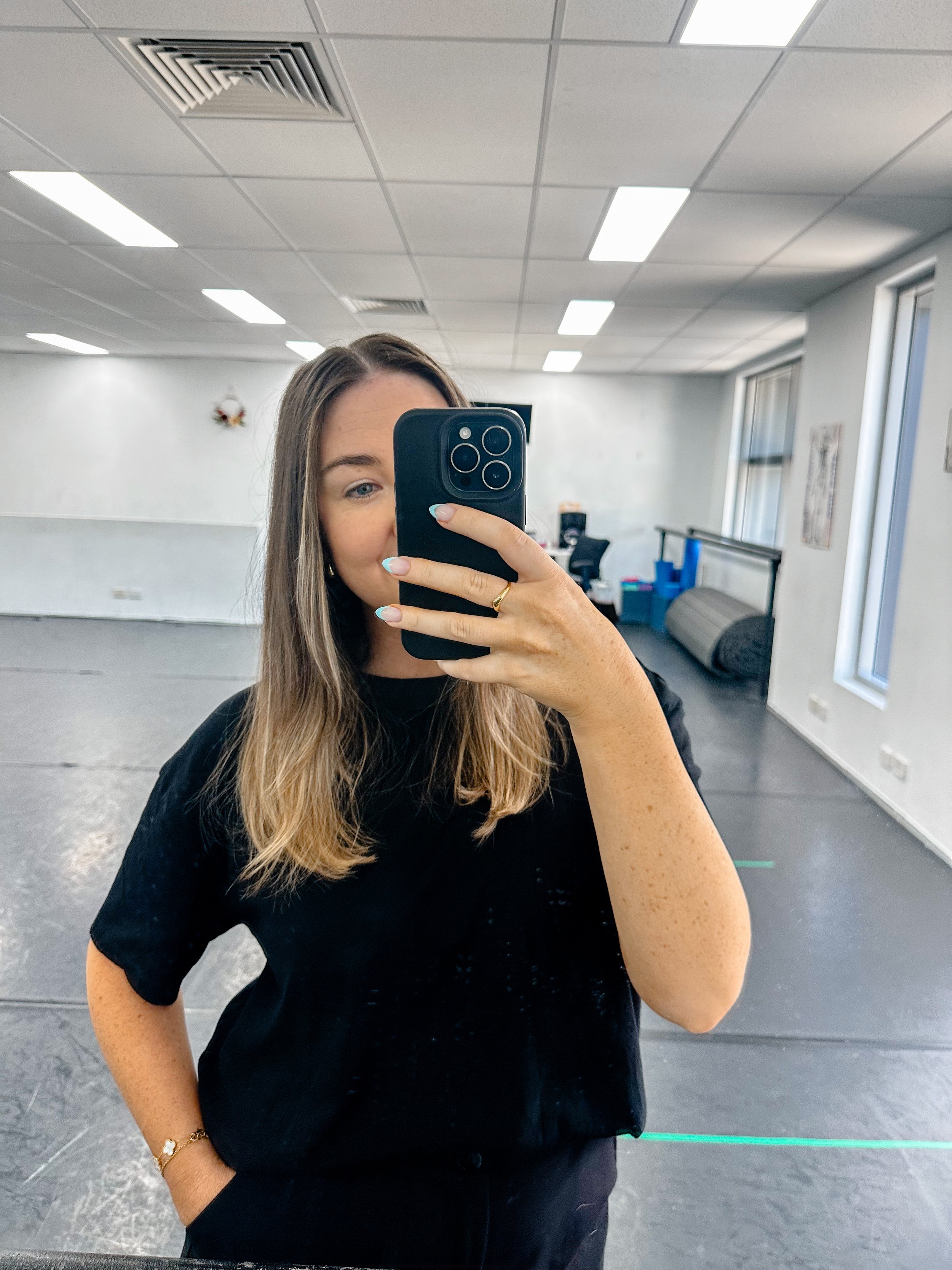 A woman with long brown hair taking a selfie in a large, empty room with white walls, ceiling lights, and windows.