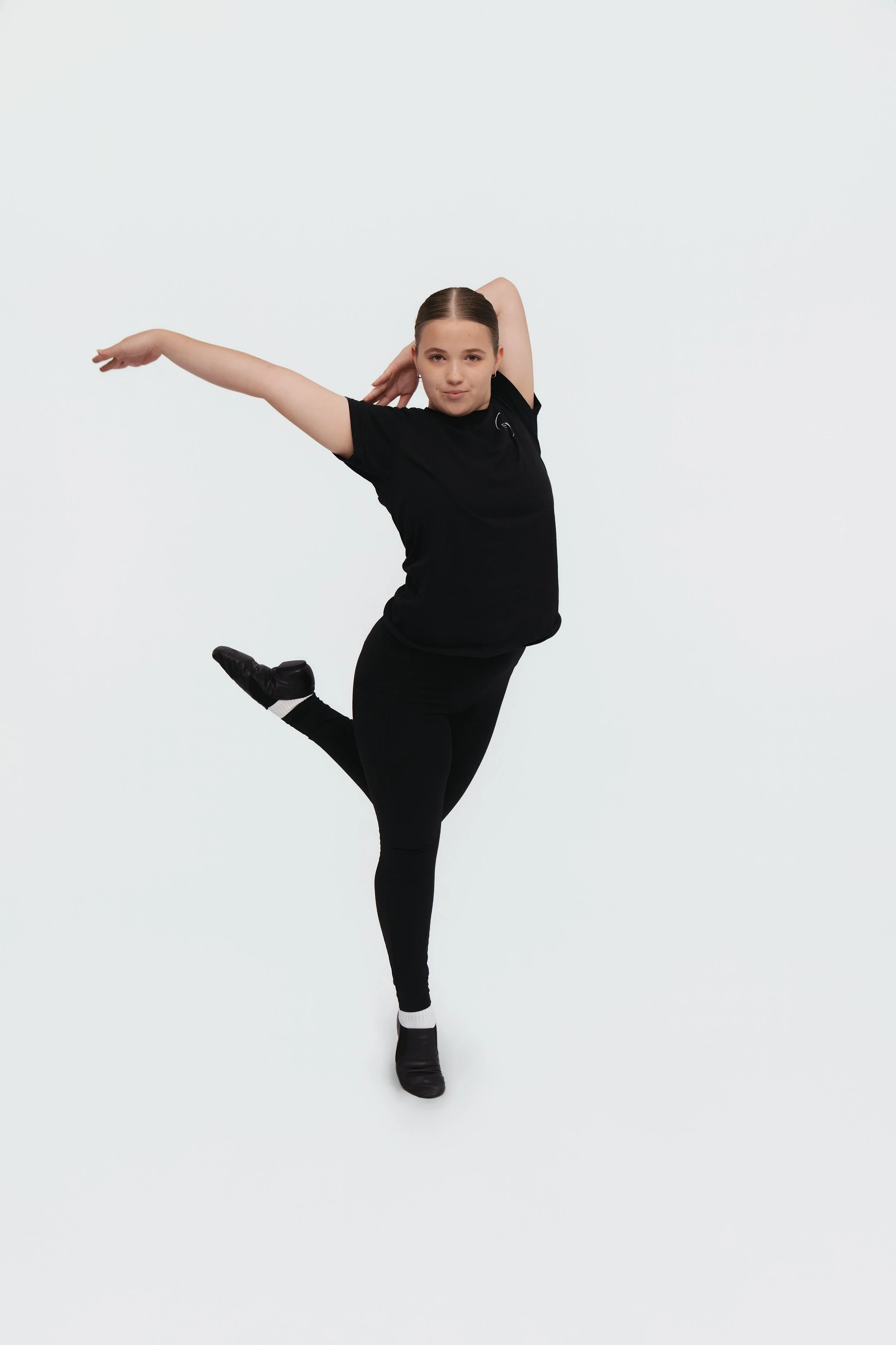 A young woman in black dance attire performing a ballet pose with one leg lifted and arms in a stylized position against a plain white background.