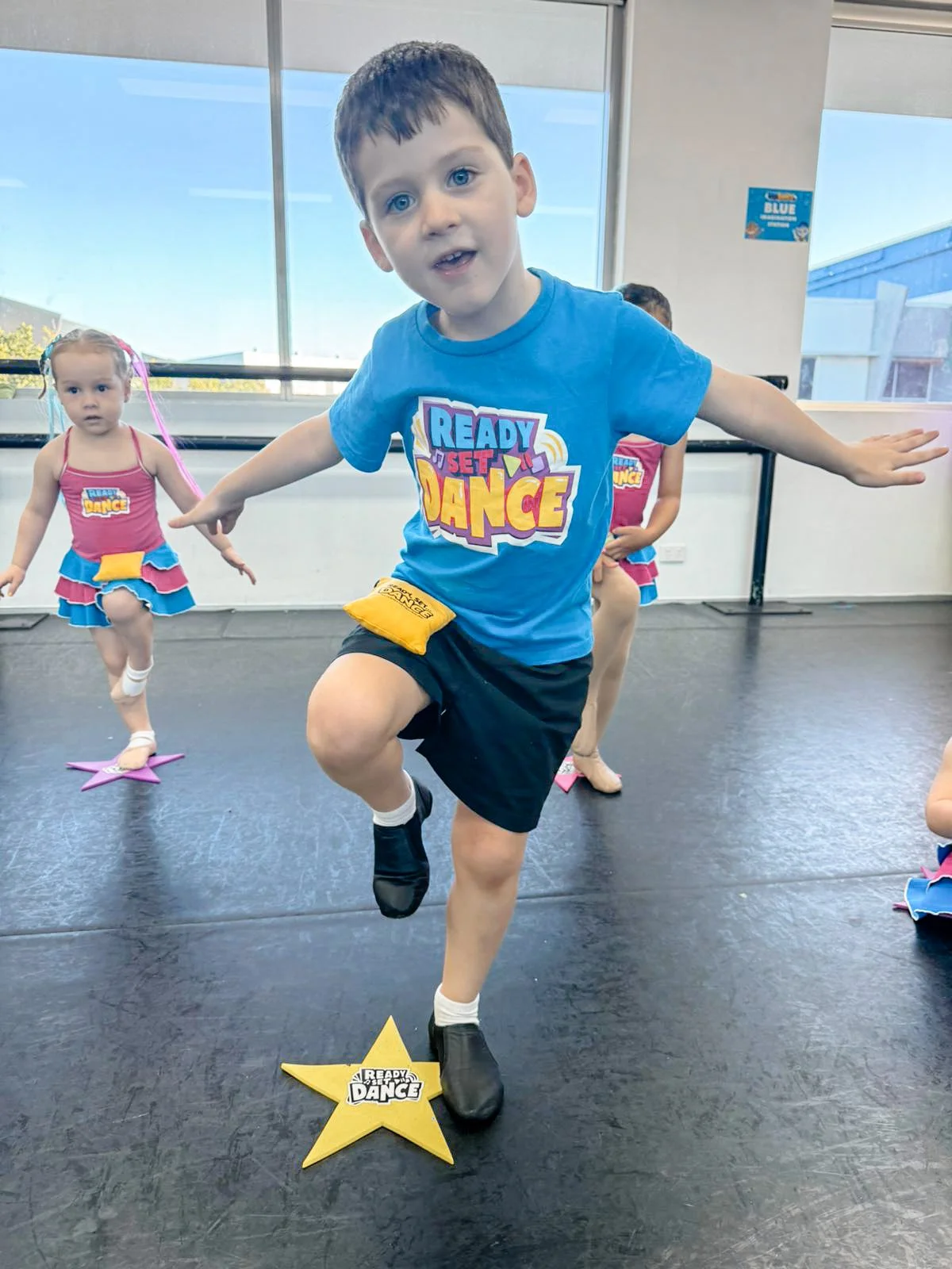 Young boy participating in a dance class, stepping on a yellow star with the text 'Ready Set Dance,' surrounded by other children in colorful dance costumes in a studio with large windows.