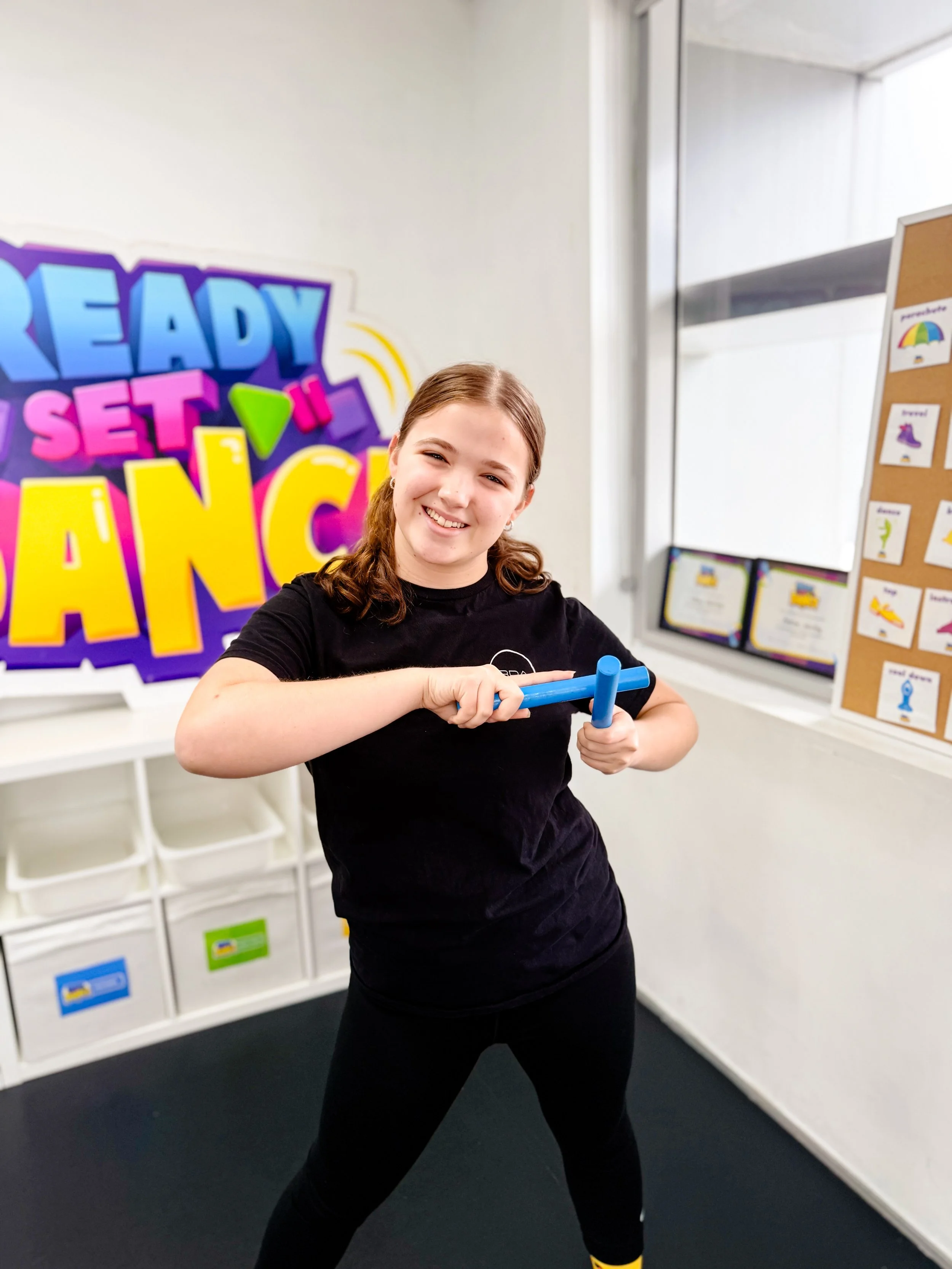 A young girl smiling and holding a blue rolling pin in a classroom or activity room with colorful signage and decorations in the background.