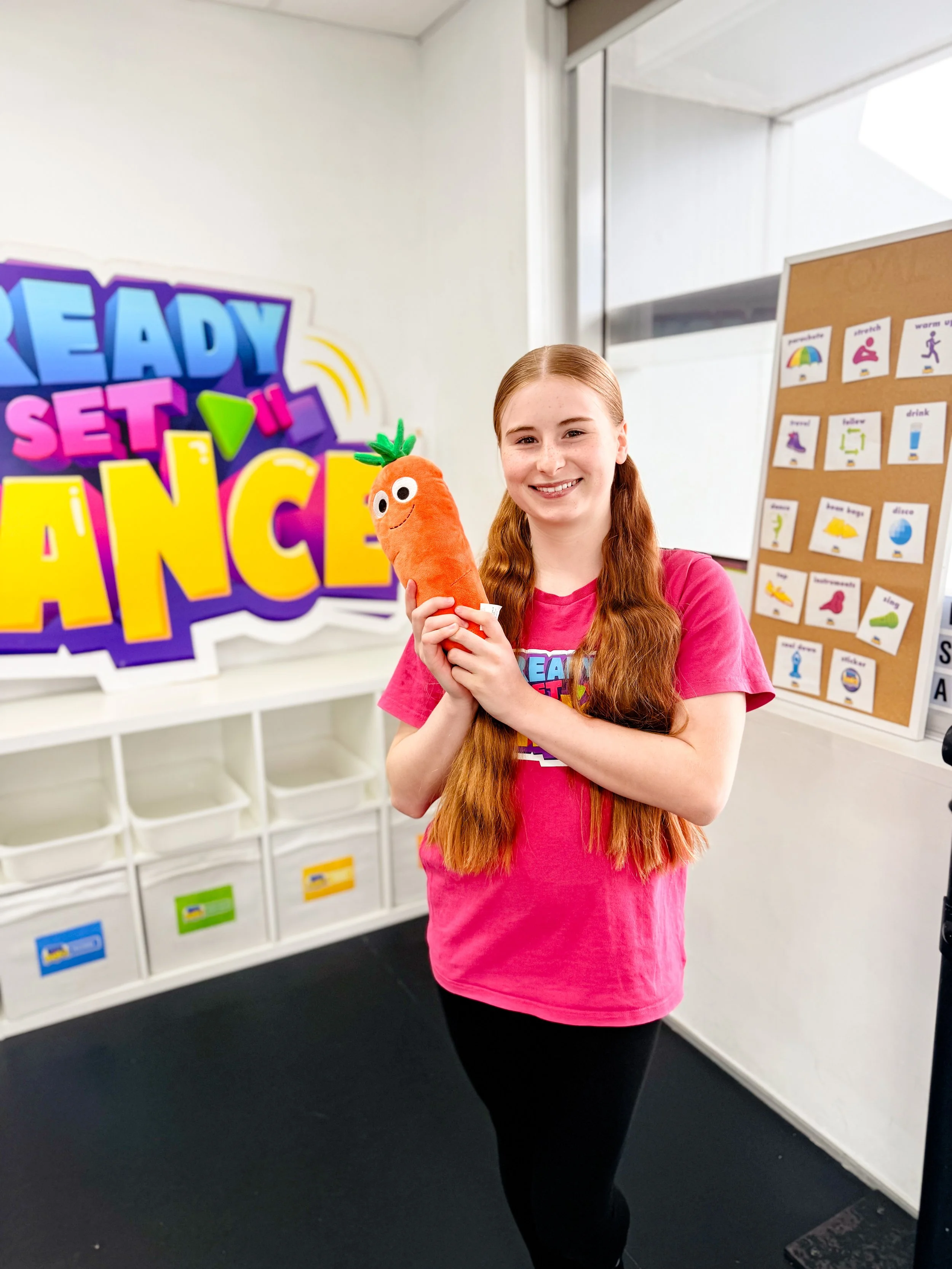 Girl with long red hair smiling and holding a plush carrot toy in a classroom with colorful signs and educational posters.