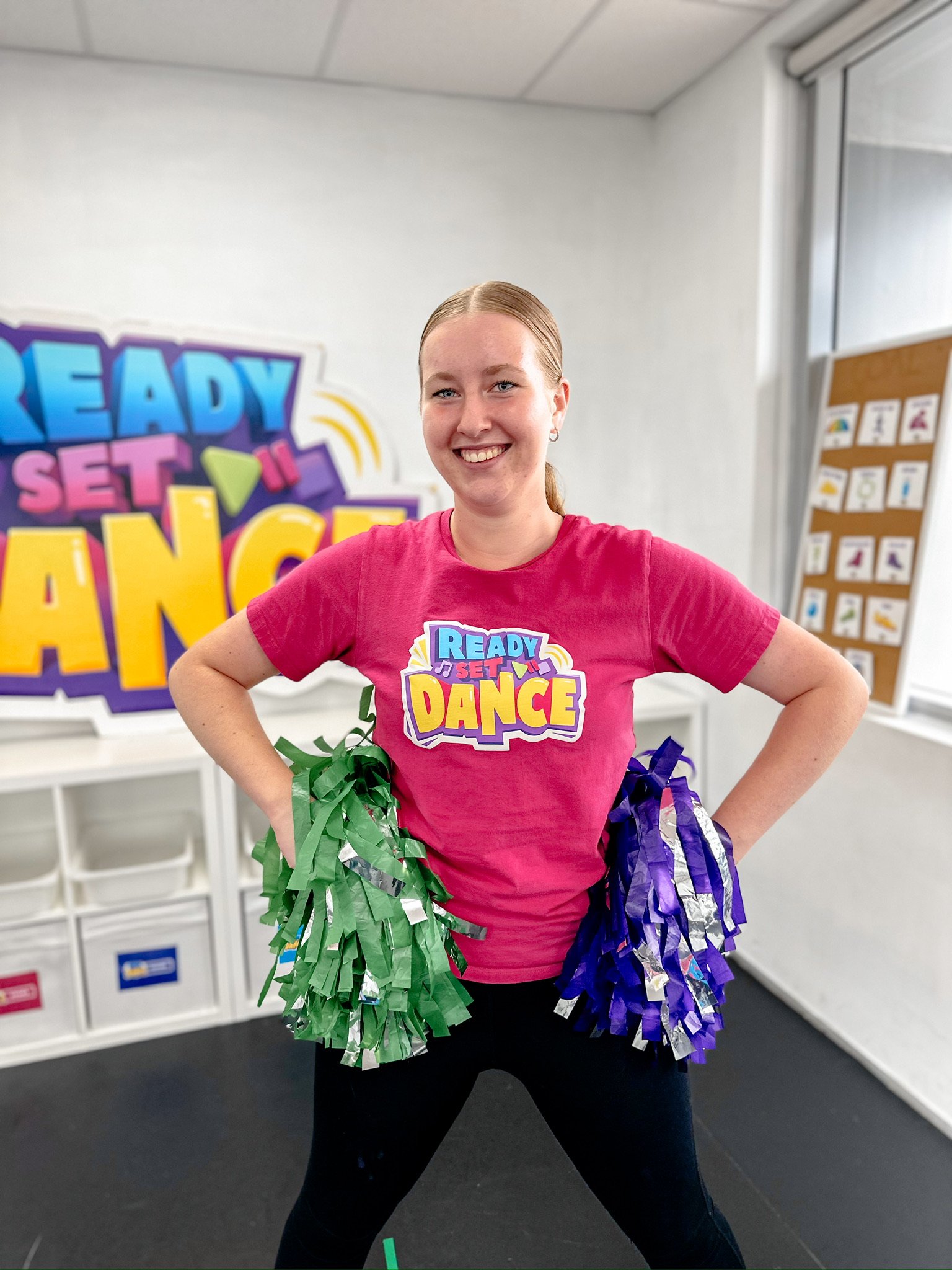 A young woman with red hair in a ponytail, wearing a pink t-shirt that says 'Ready Set Dance,' holding green and purple pom-poms, smiling and standing in a decorated room.