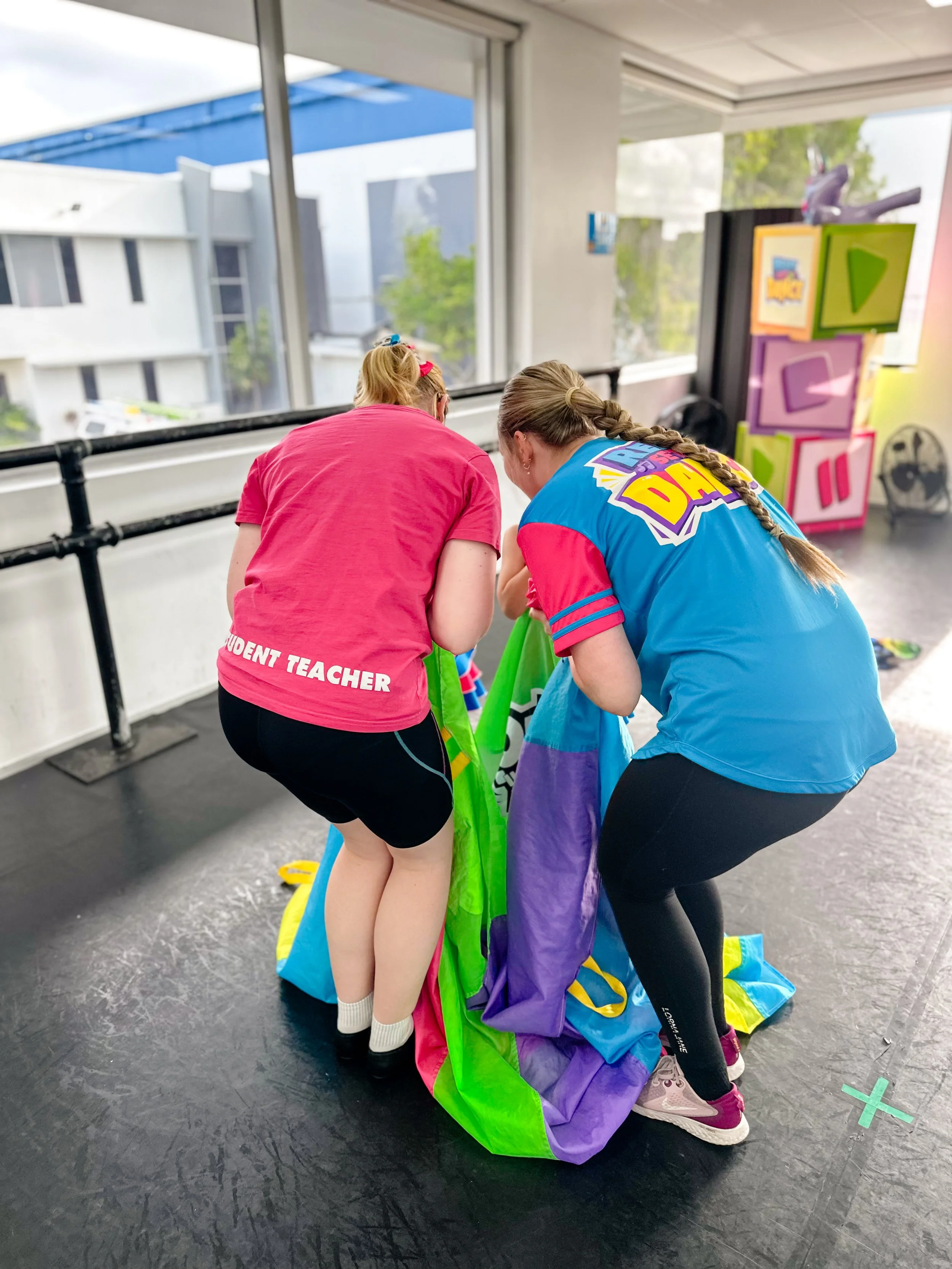 Two women, one wearing a pink shirt with 'Student Teacher' on the back and the other in a blue and pink shirt, are folding or organizing a colorful parachute on a stage or performance area.
