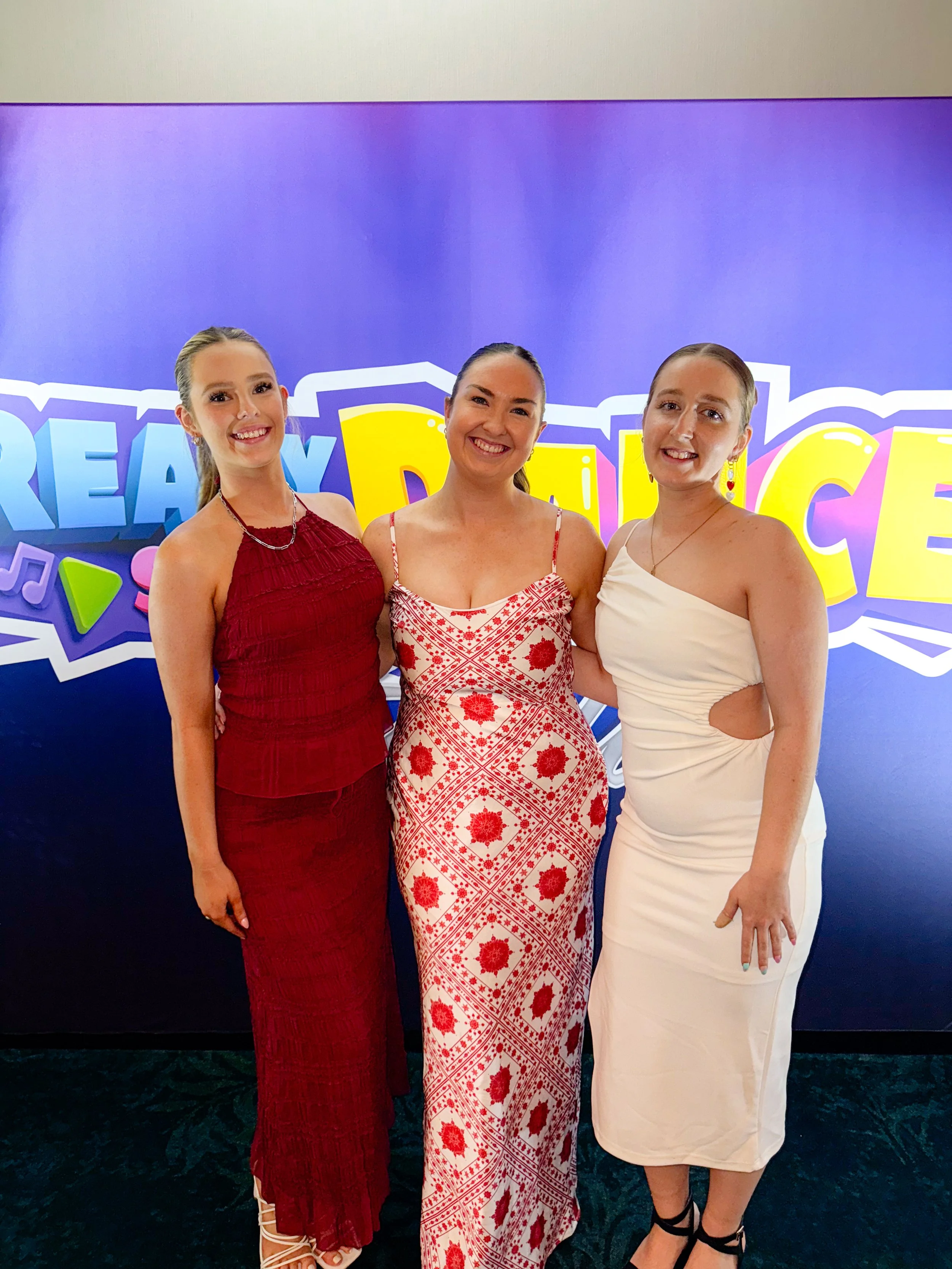 Three women in formal dresses standing in front of a colorful backdrop with the words "Dream Dance".
