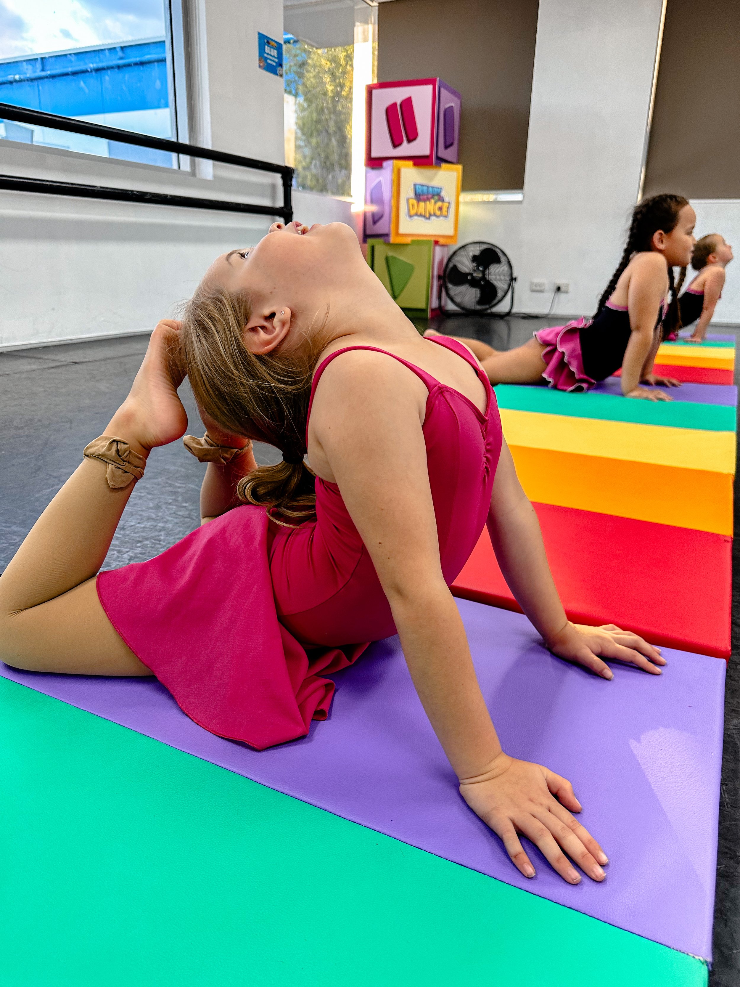 Young girl in a pink dance outfit performing a yoga pose on colorful mats in a dance studio.