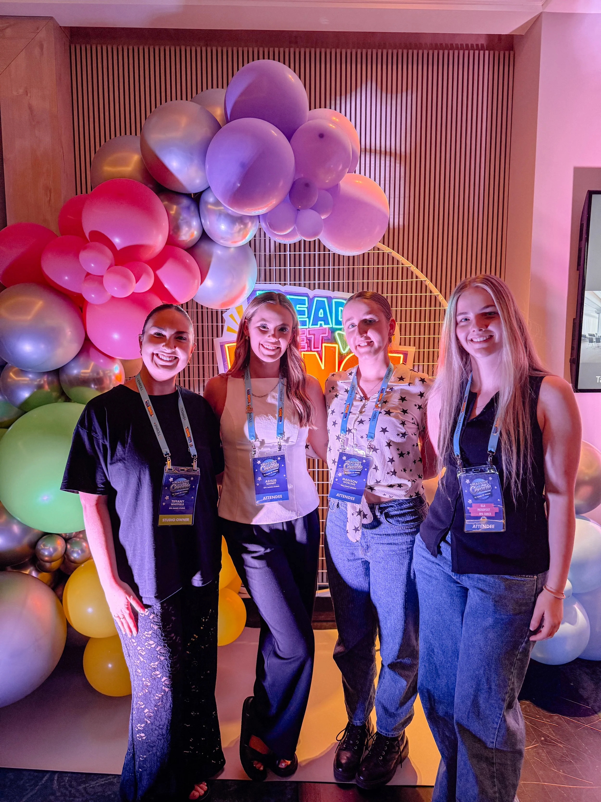 Four women standing together, smiling at an event with a colorful balloon arrangement and a neon sign in the background.