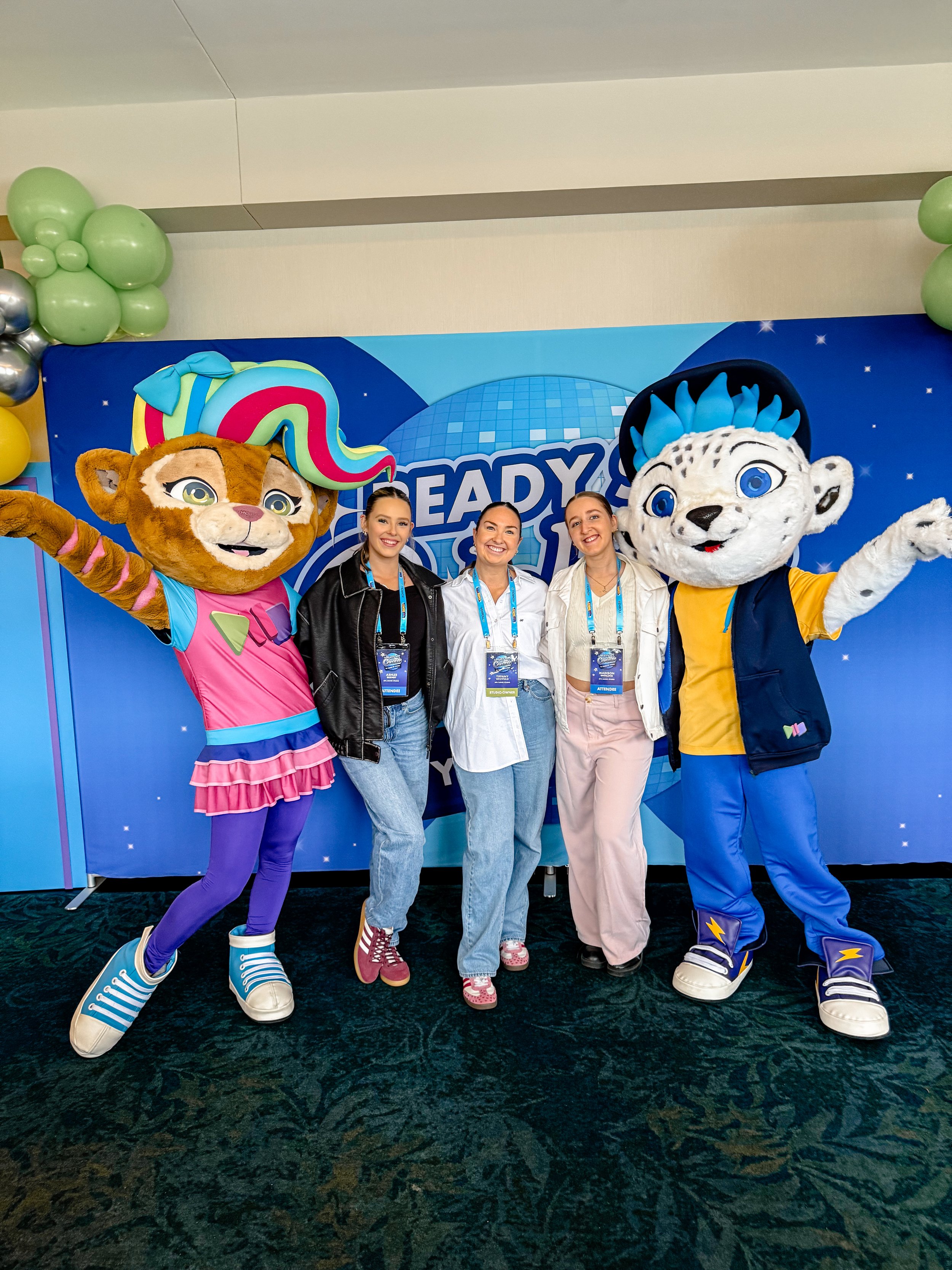 Three women in casual clothing standing with two costumed characters in front of a blue backdrop with balloons, at an event called 'READY'.