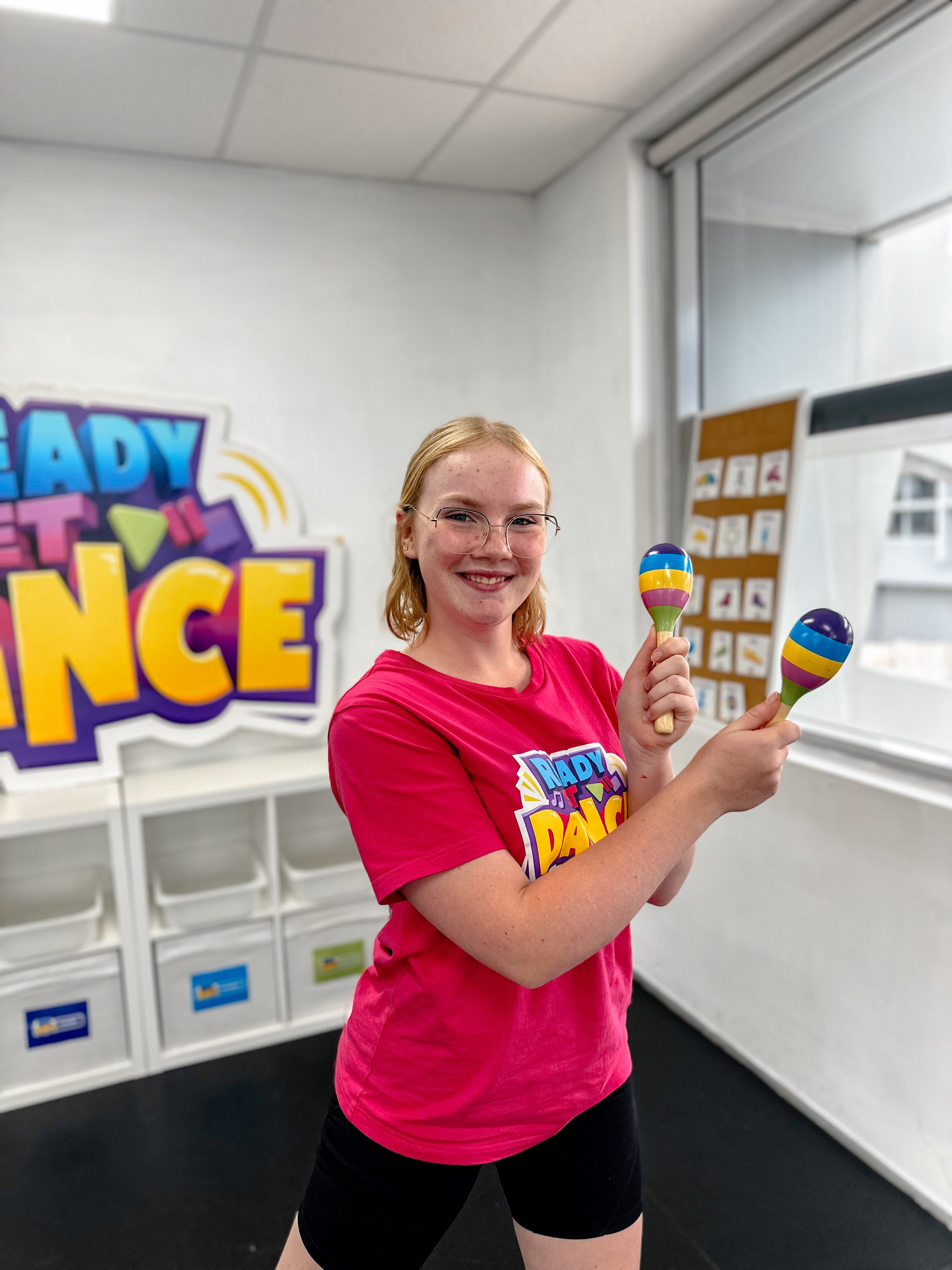 A young girl with red hair, glasses, and freckles smiling and holding maracas in a dance studio. She is wearing a pink T-shirt with a colorful logo that says 'Ready to Dance.' There is a large sign in the background with the same logo and a bulletin 