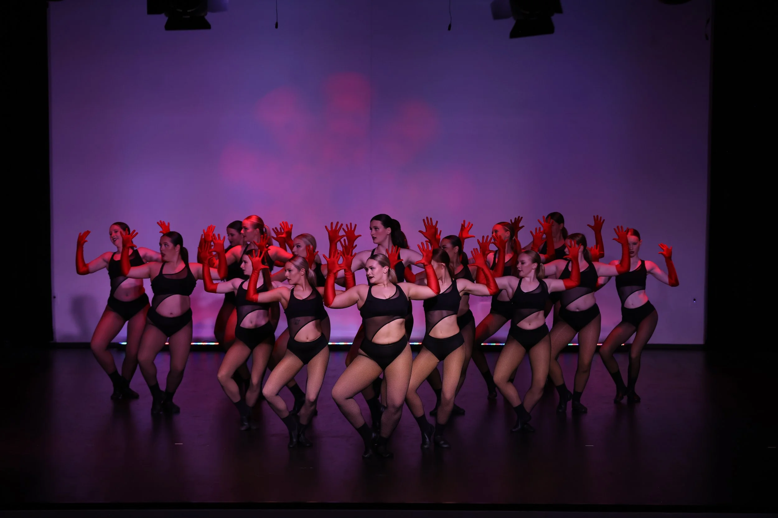A group of female dancers performing on stage in black costumes with fishnet tights and red gloves. They are standing in a formation under stage lighting with a purple backdrop.