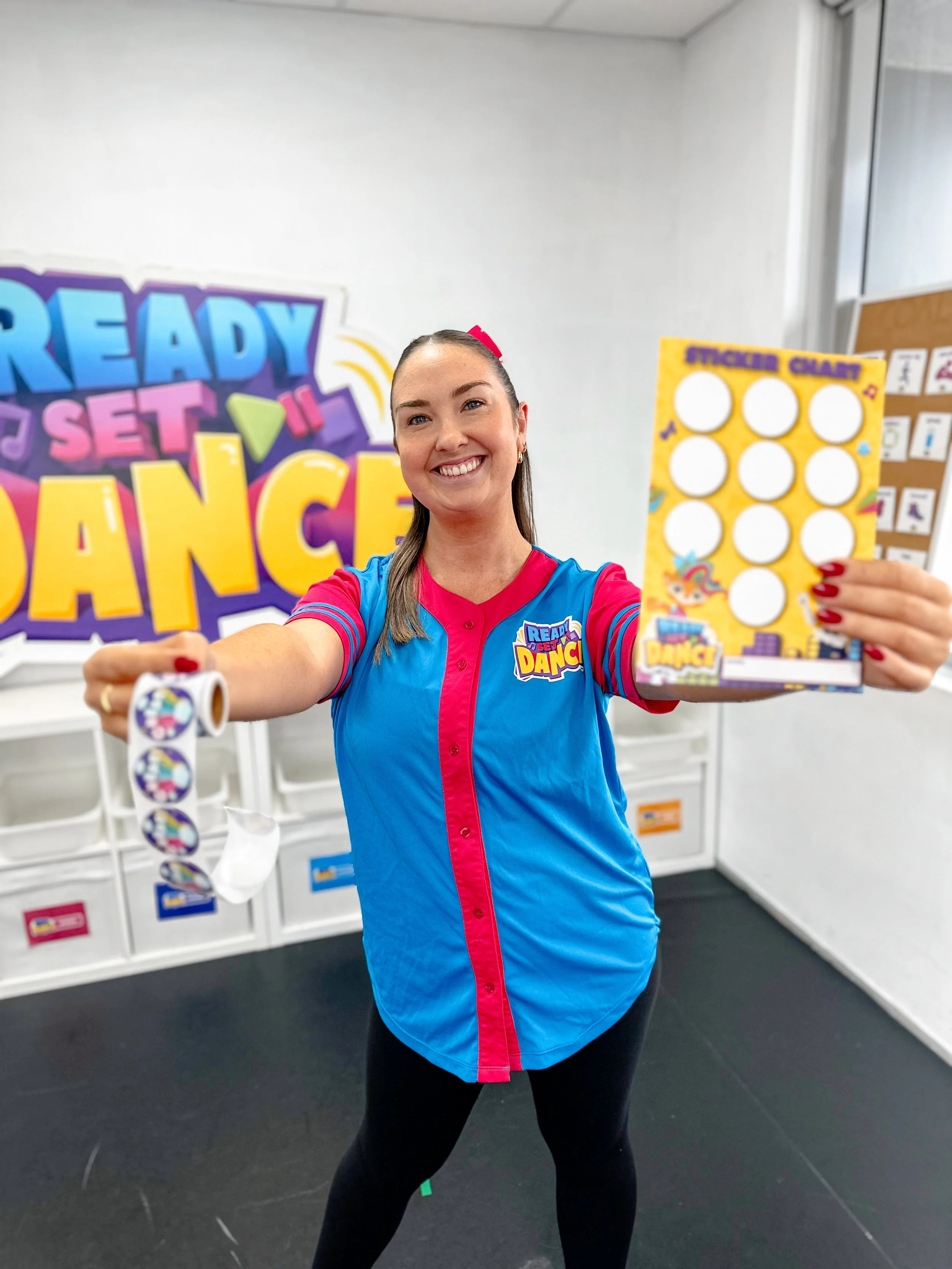 A woman in a colorful shirt holding bingo stickers and a roll of stickers in a classroom with a sign that reads 'Ready, Set, Dance' in the background.