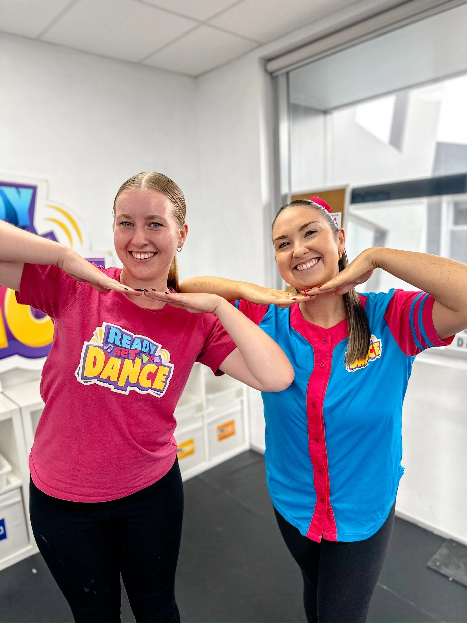 Two women in colorful shirts smiling and posing inside a room, with one in a pink shirt and the other in a blue and pink shirt, both making a fun gesture with their arms.