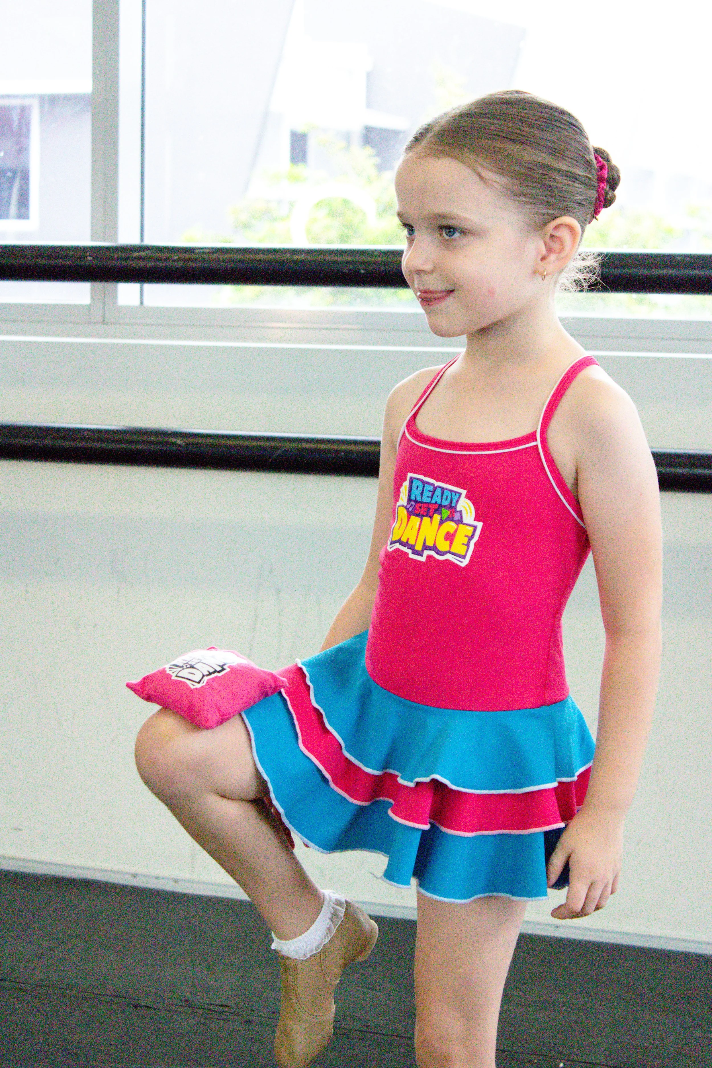 A young girl in a pink and blue dance costume with a 'Ready Set Dance' logo, practicing ballet in a dance studio.