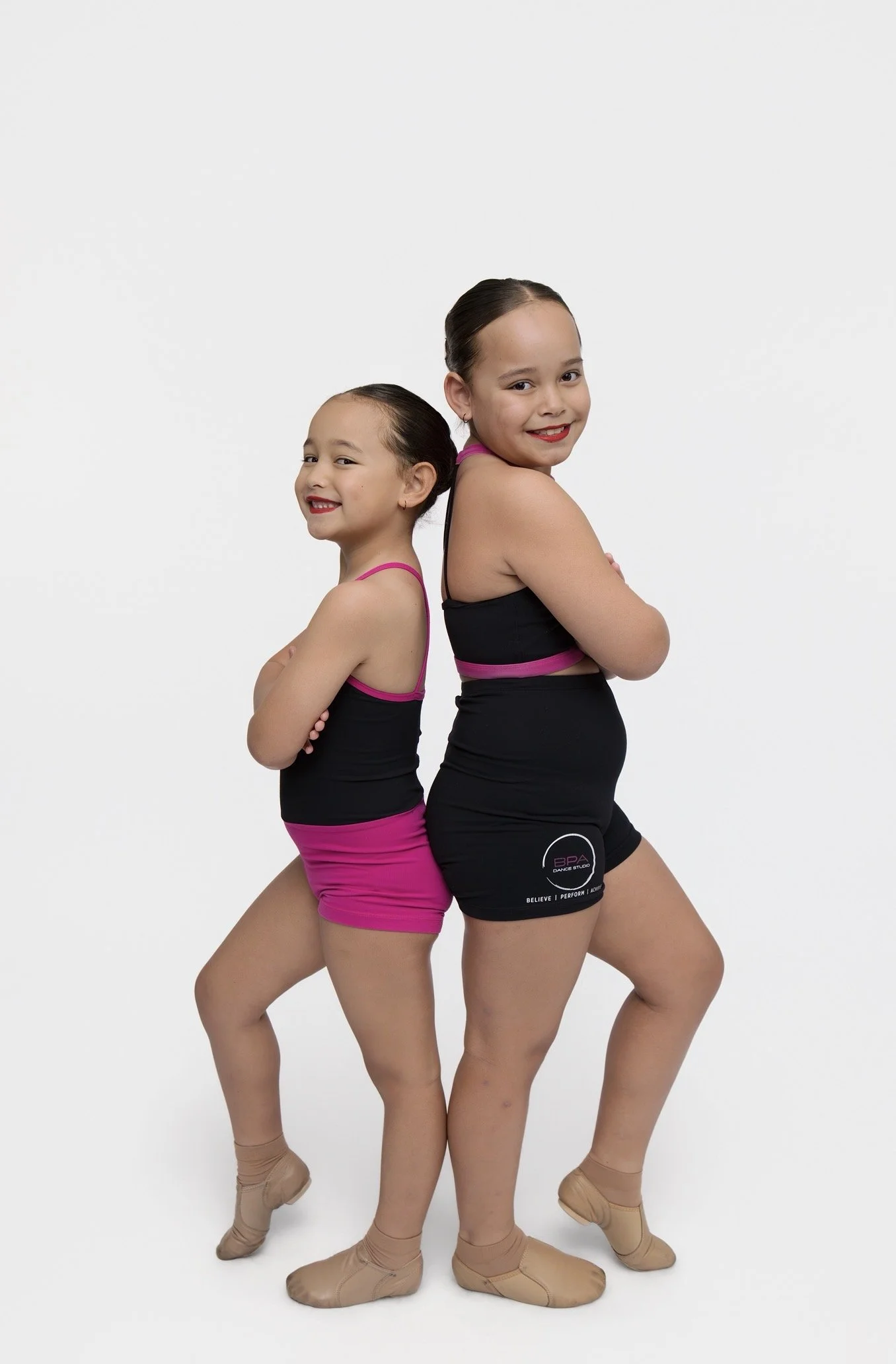 Two young girls in dance costumes standing back to back with crossed arms, smiling. They wear black and pink dance outfits, tan ballet shoes, and have their hair slicked back.