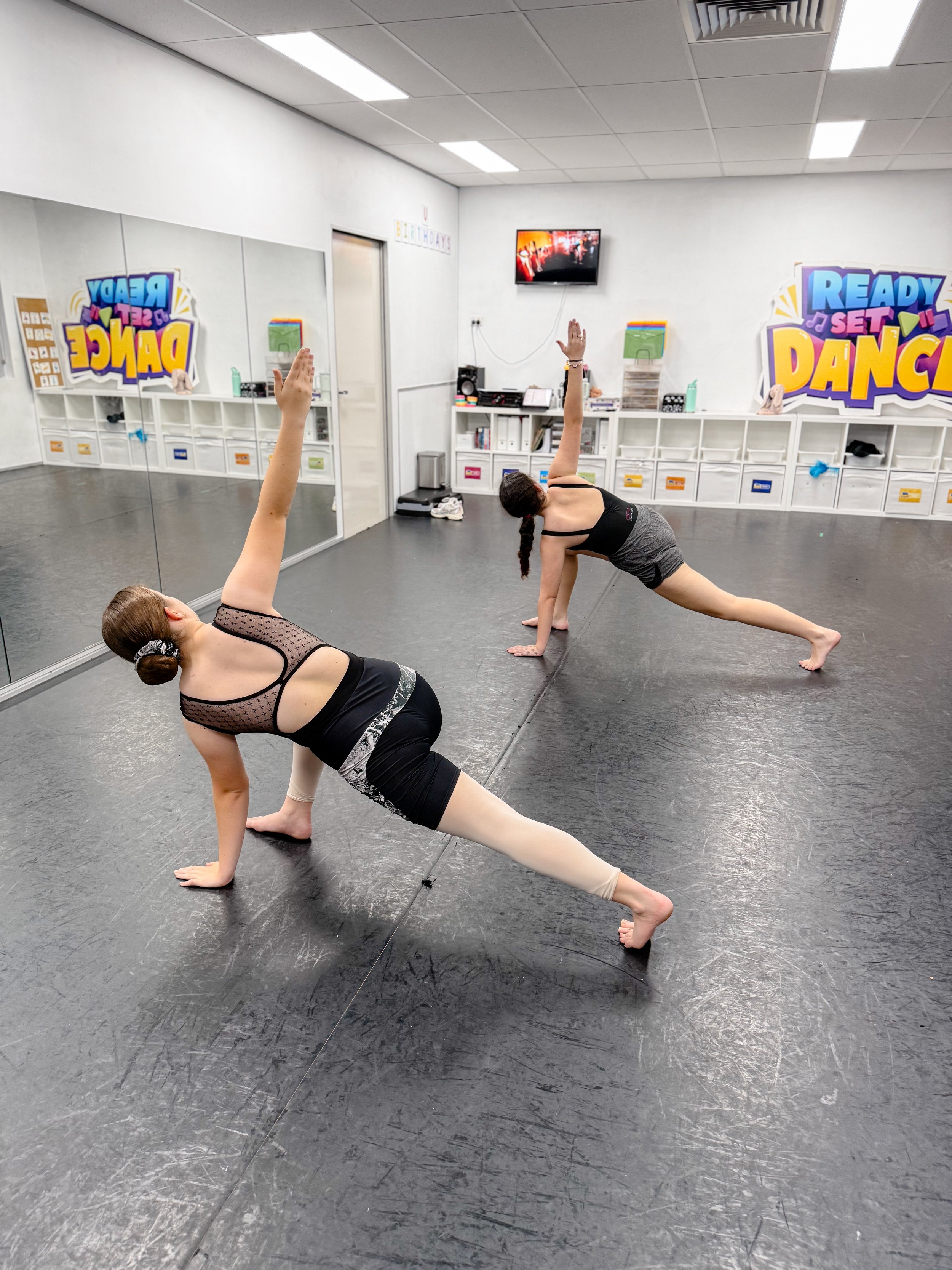 Two young girls practicing yoga in a dance studio with colorful signs on the wall reading 'Ready Set Dance' and 'Ready Set Dance'. They are in a side plank pose with one arm raised, wearing athletic clothing.