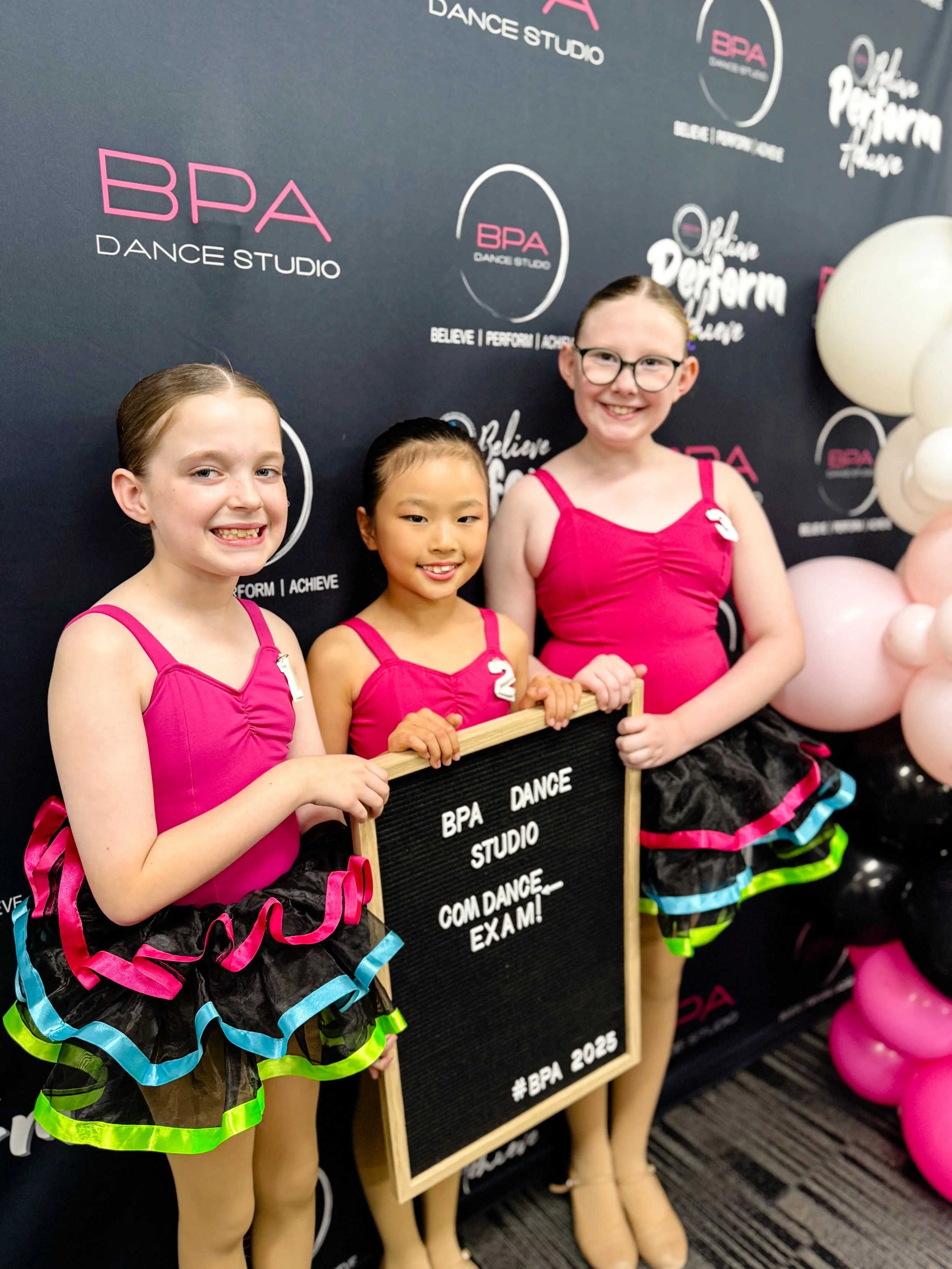 Three young girls in colorful dance costumes standing in front of a black backdrop with pink and white logos, holding a black sign that reads 'BPA DANCE STUDIO COME DANCE EXAM! #BPA 2025'. They are smiling and posing for a photo.