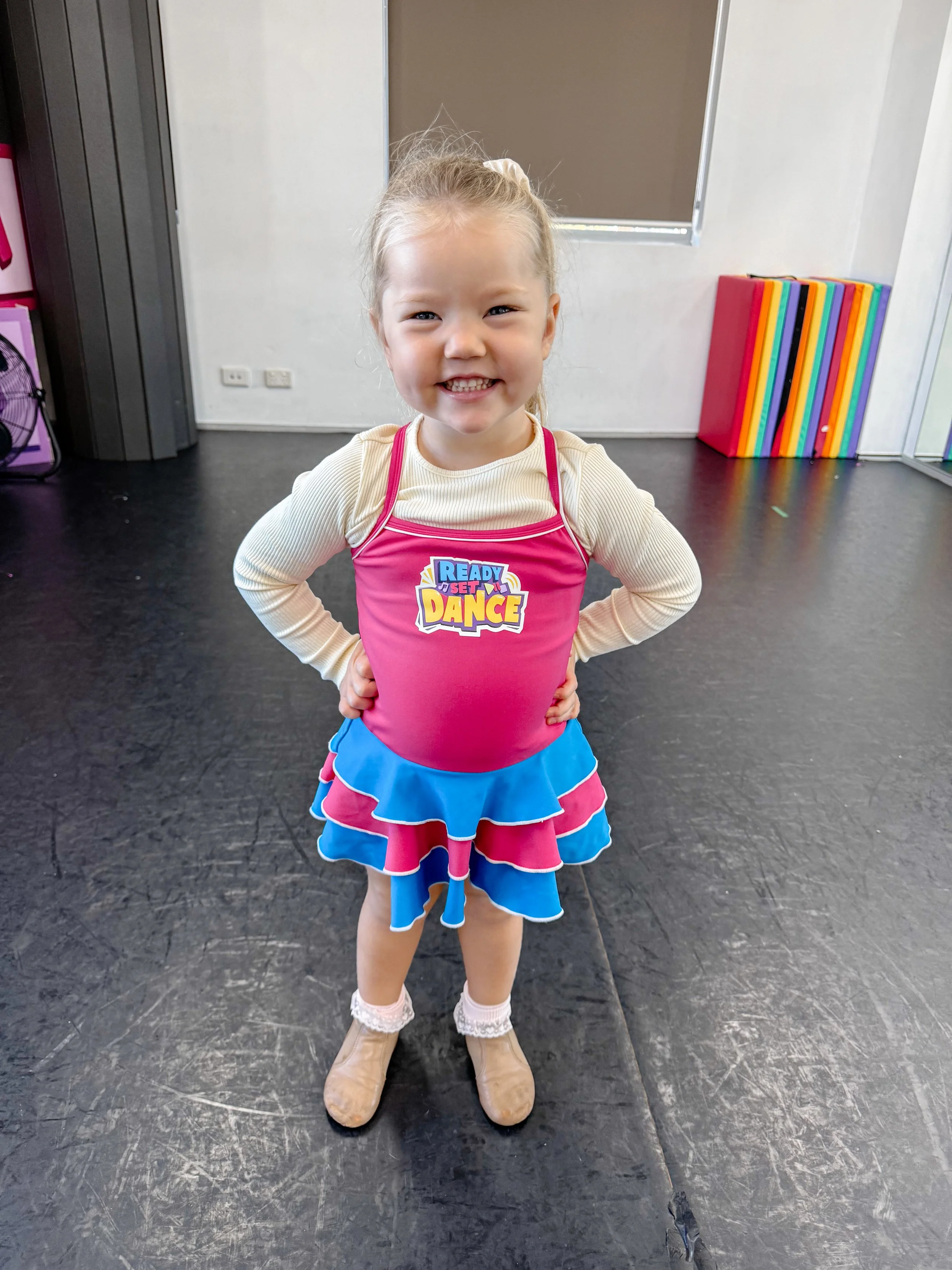A young girl smiling with hands on her hips, wearing a pink dance costume with a blue and pink layered skirt, beige boots, and a beige long-sleeve shirt, standing in a dance studio.