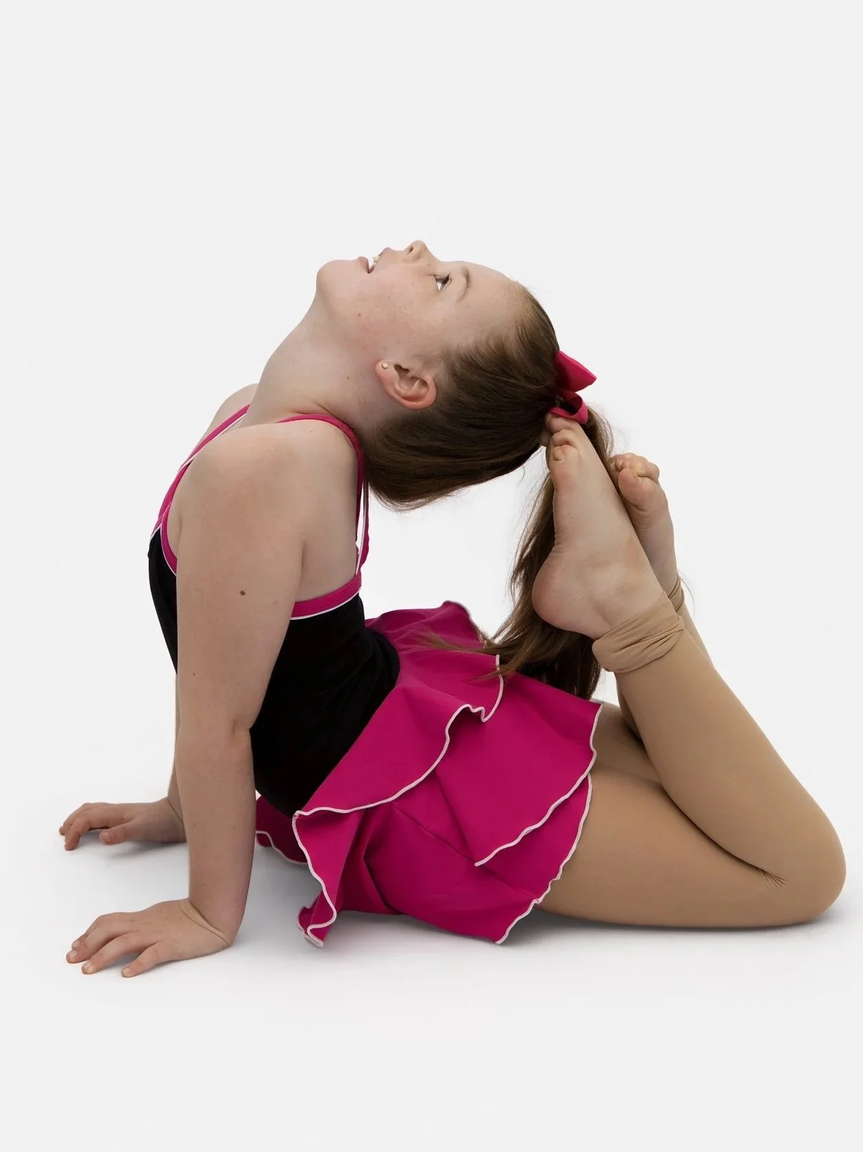 A young girl dressed as a ballerina practicing a yoga pose where she bends backward and holds her foot with her hand, wearing a pink and black leotard with a pink skirt and a pink bow in her hair.