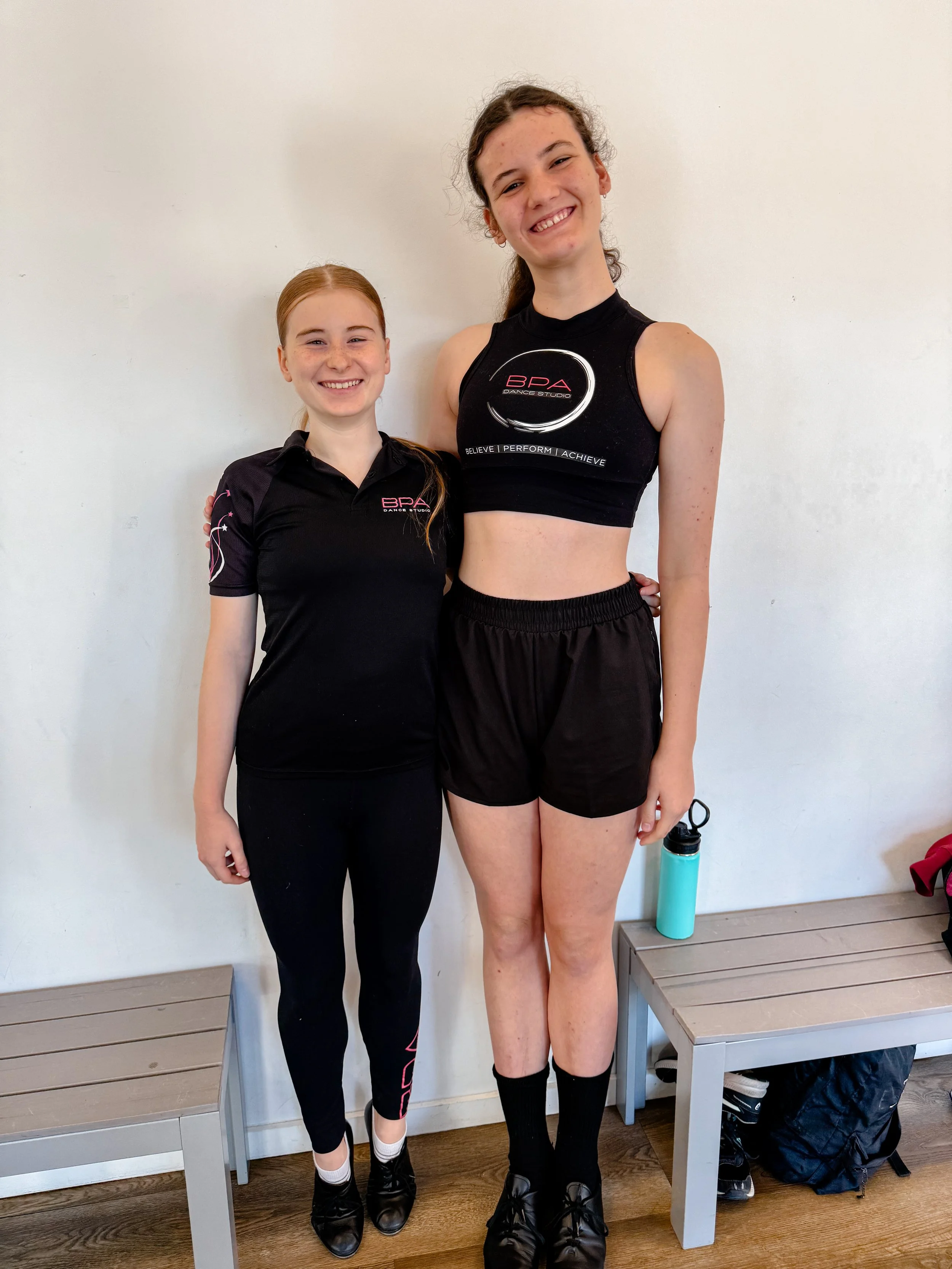 Two young female dancers standing side by side indoors, smiling. The smaller girl is dressed in a black dance uniform with pink accents, and the taller girl is wearing a black crop top with a logo and black shorts, holding a water bottle.