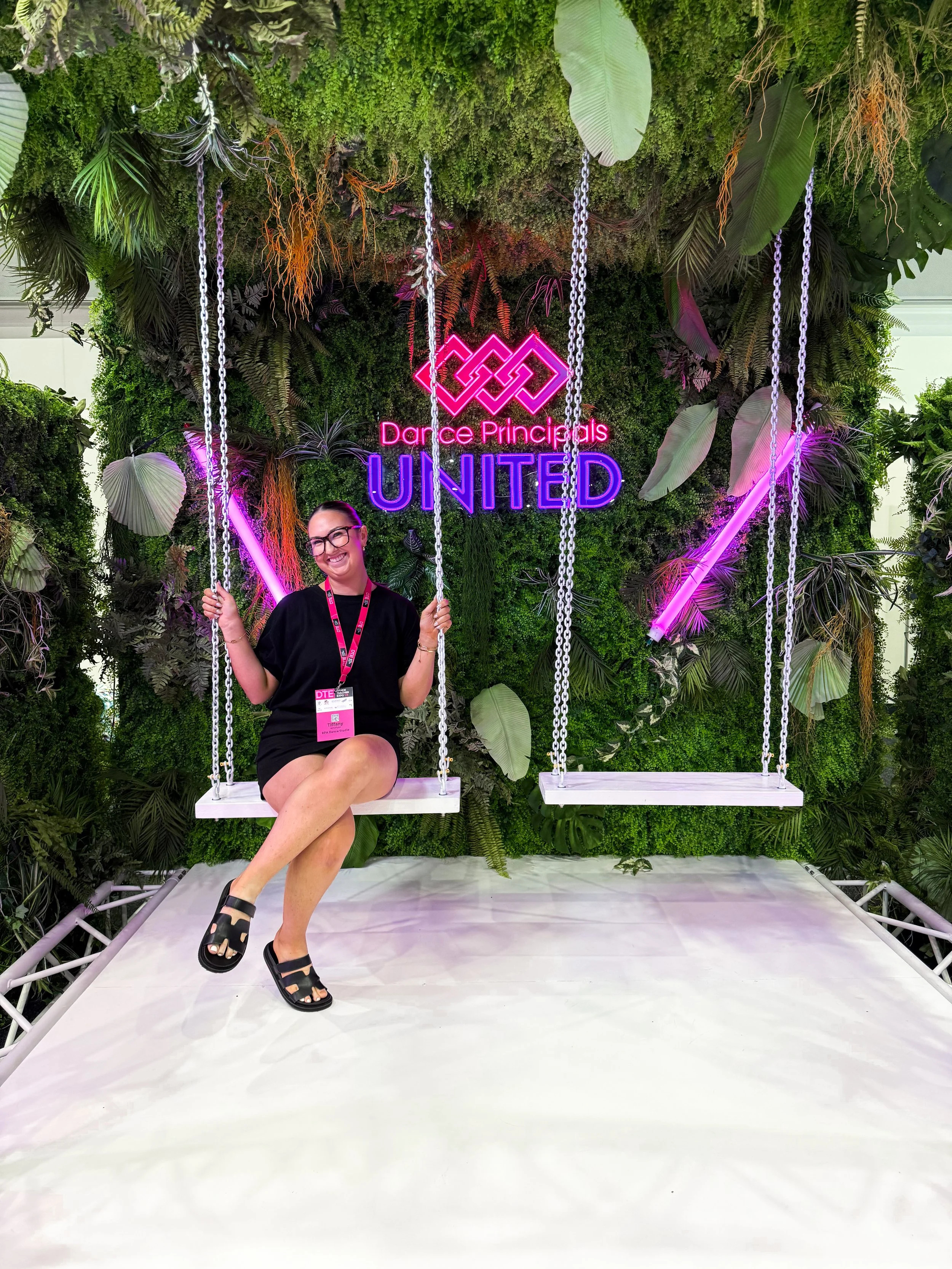 A woman sitting on a swing in front of a lush green moss and plant wall with a neon sign that reads 'Dance Principals UNITED' in pink and purple lights.