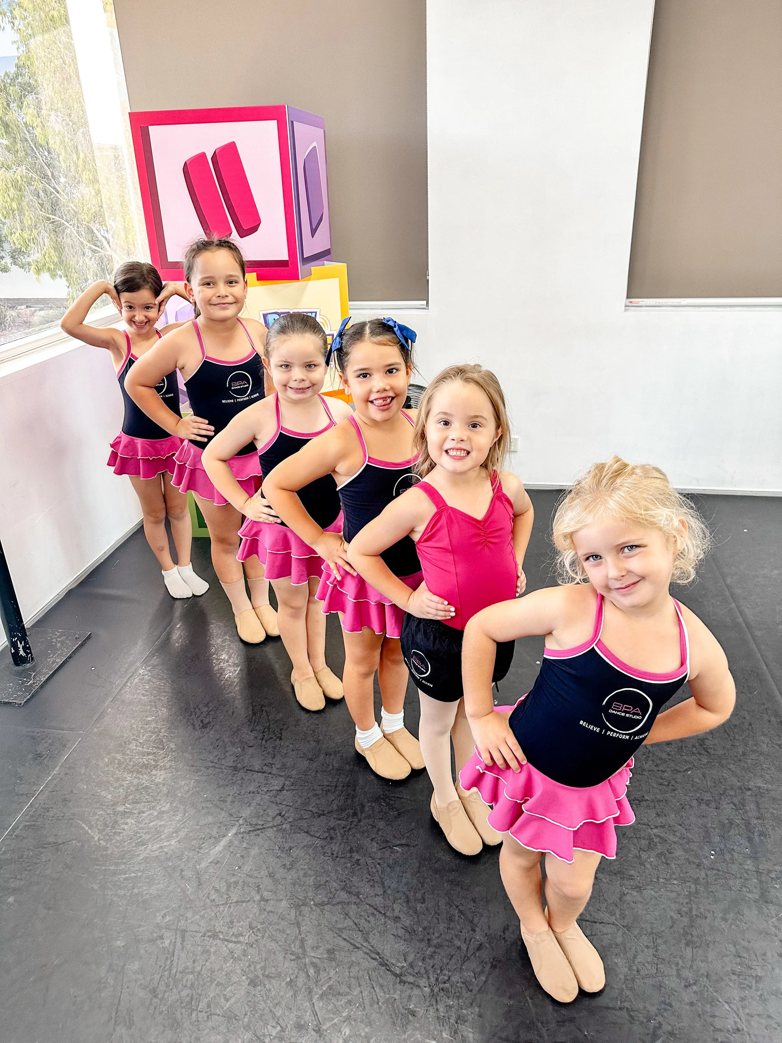 Six young girls in ballet costumes standing in a line, smiling, inside a dance studio with a large window and a colorful cube with ballet shoes illustration in the background.