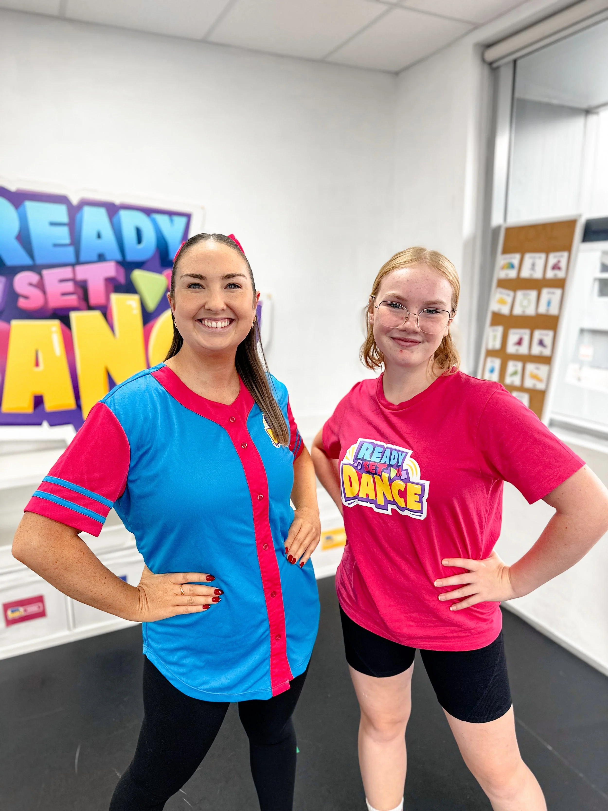 Two women posing in a room with a colorful sign that reads 'Ready Set Dance' in the background; the woman on the left is wearing a blue and pink dance shirt, and the woman on the right is wearing a pink t-shirt with the same logo.