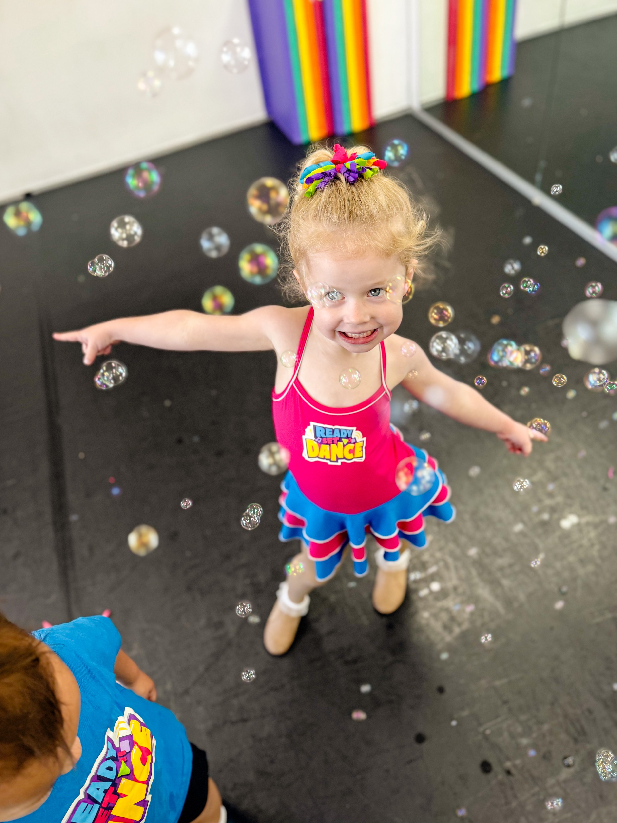 A young girl in a bright pink dance outfit with a colorful skirt and a badge that says 'Ready Set Dance' stands in the middle of a room with floating bubbles, smiling and pointing to her right.