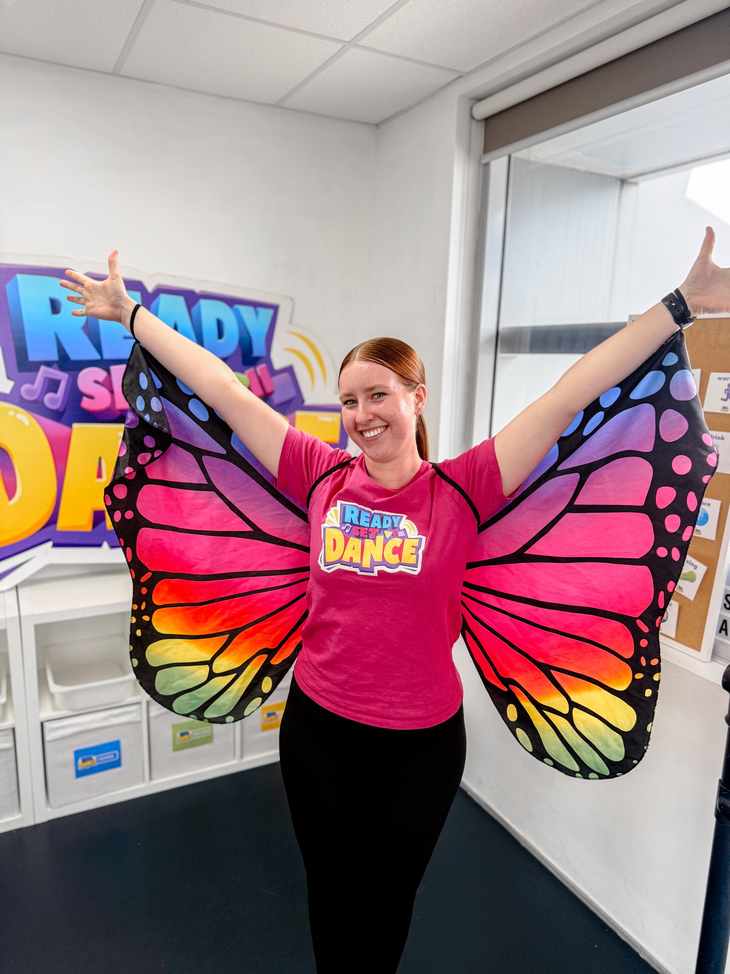 A woman with red hair wearing a pink shirt with a 'Ready Set Dance' logo, black pants, and large butterfly wings with pink, orange, yellow, green, blue, and purple colors.