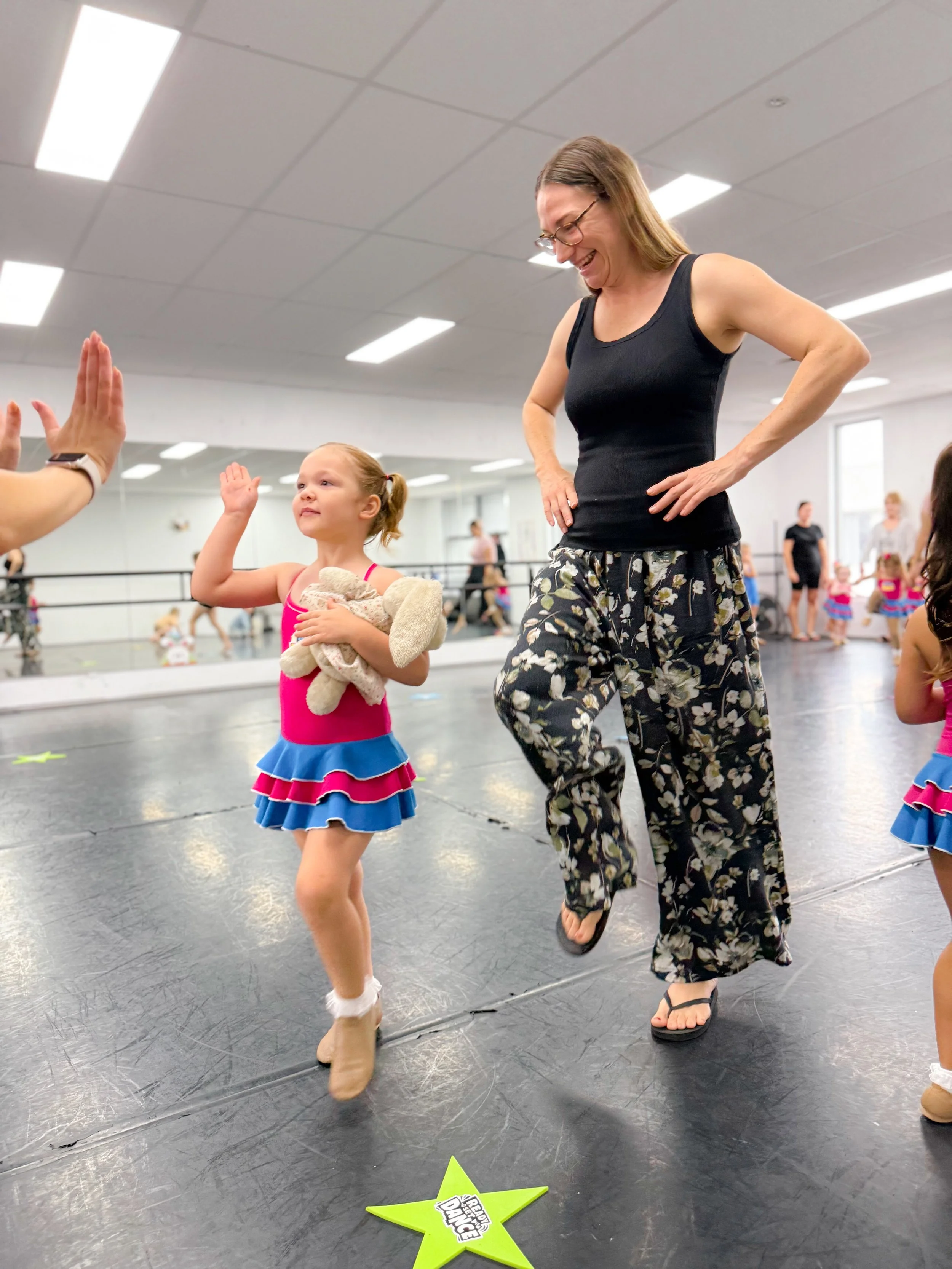 A dance instructor, wearing glasses, a black tank top, and floral pants, smiles while giving a high-five to a young girl in a pink and blue dance costume in a dance studio. Other children and adults are in the background.